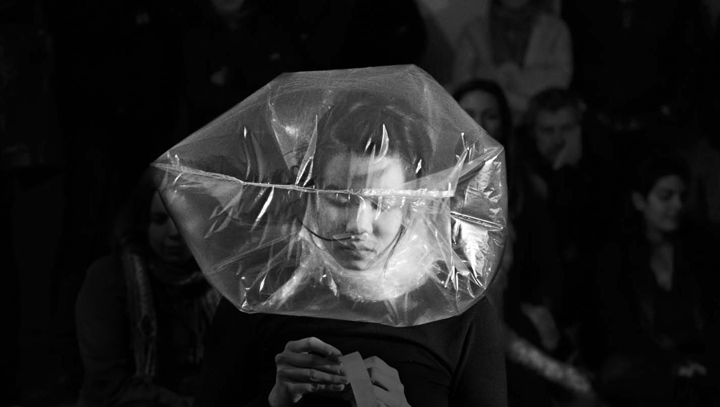 An inflated plastic bag covers the head of the artist. The artist’s face looking down her hands is visible through this translucent bag. Many people seated behind her are watching this performance. 