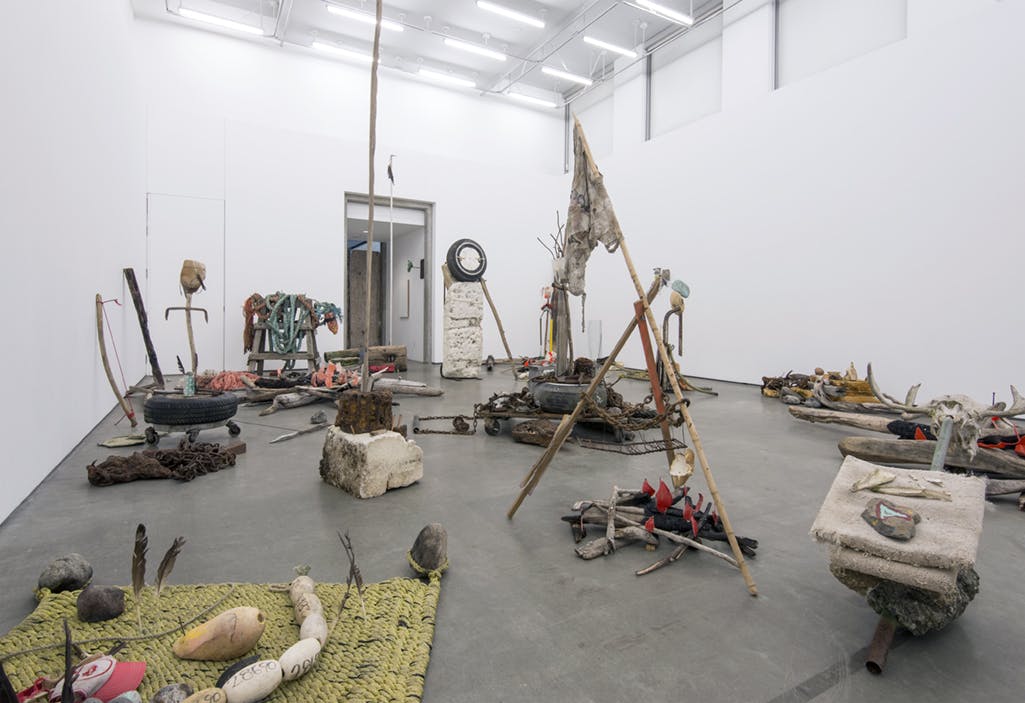 Large sculptures of found materials fill a gallery space. The overall installation mimics a camping location. Two fake bonfires, ropes, and other wooded objects are visible in this image.