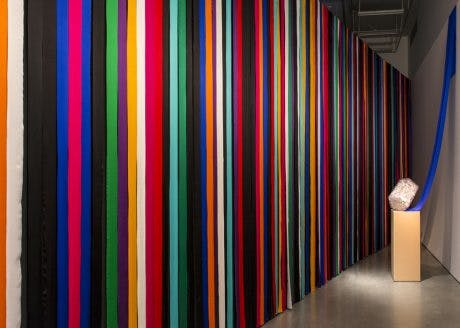 A striped multicoloured curtain hangs in a gallery. Installed in front of the curtain is a clear, block-like sculpture of encased ceramic pieces on a plinth. 