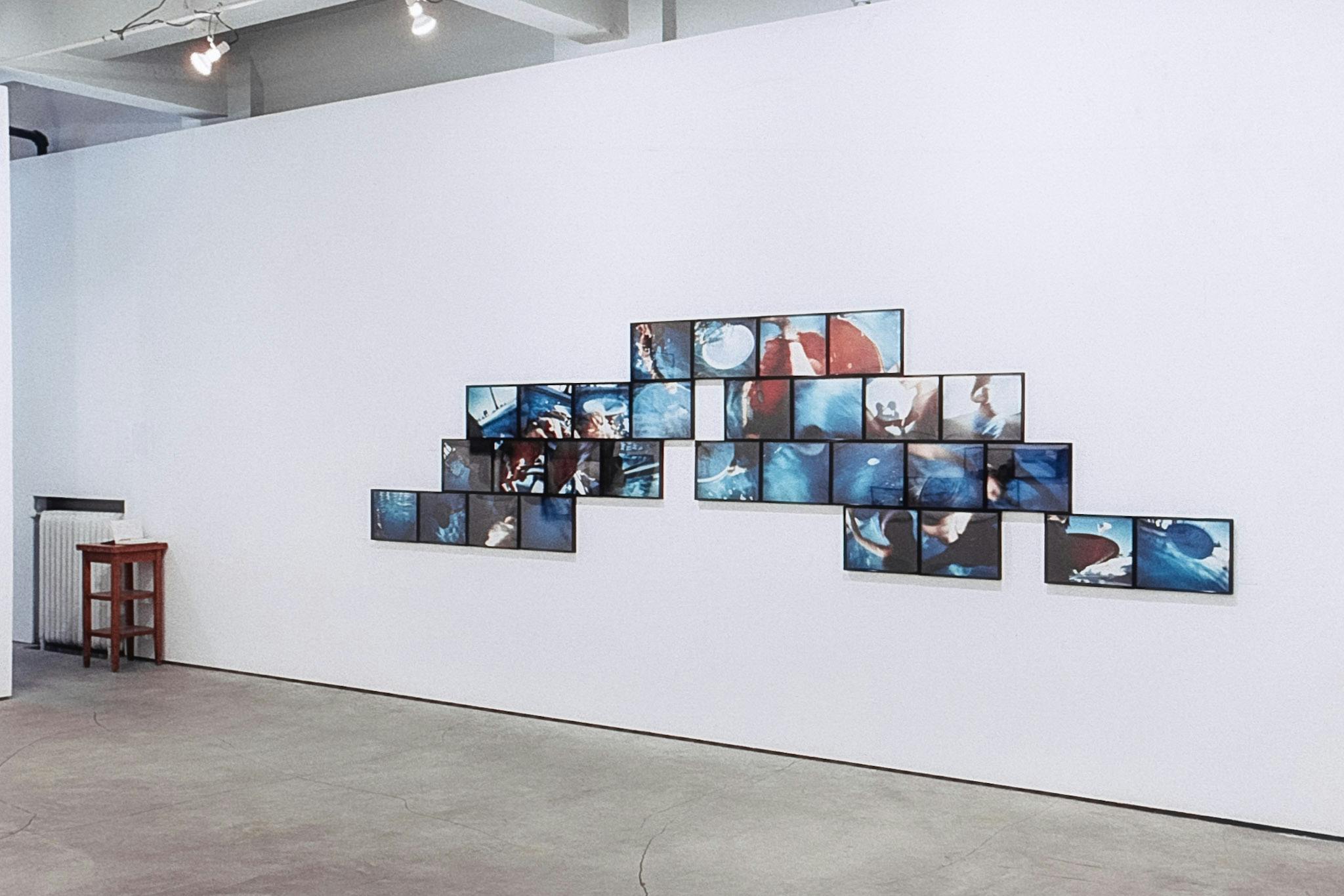On the wall of a gallery, there are 29 cibachrome photos arranged in a pyramid-like way. They show small glimpses of people and water.  In the corner of the photo, there is a small wooden side table.
