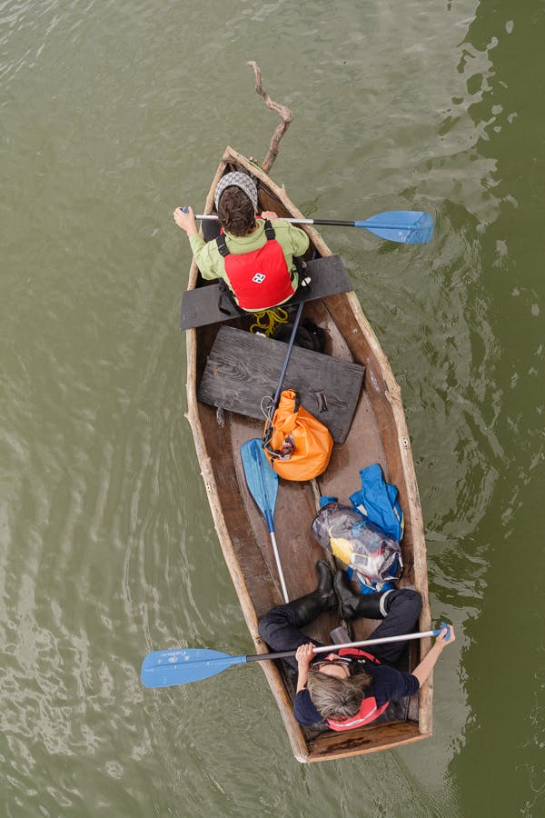 This is an aerial view of the artist, Marie Lorenz and her company riding on a wooden boat. They puddle to move through the water. Some large bags are placed in between where those people sit.