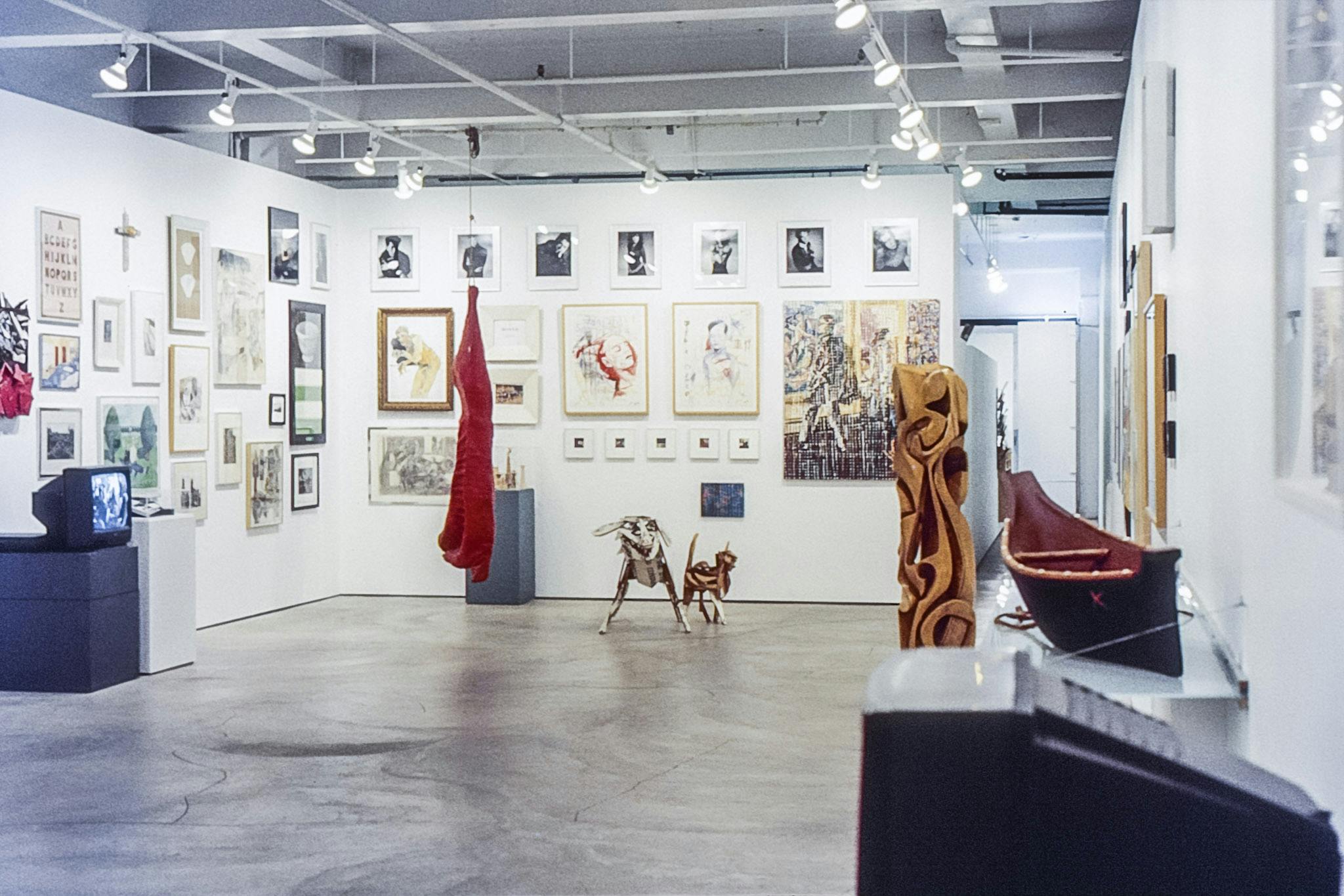 A photo of various artworks installed close to each other in the gallery. On the floor, there are two TVs playing video works, one canoe, a wooden abstract statue, and a dog and a cat made with metal.