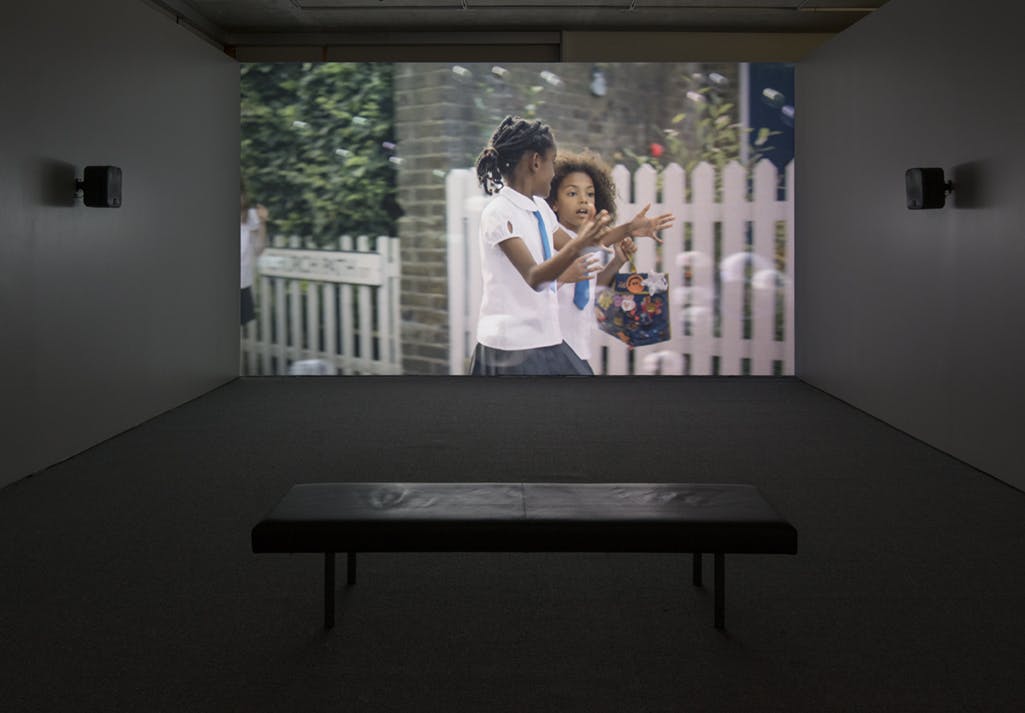 A single-channel video is projected onto a gallery wall. The video shows two children in school uniforms walking down a street, along a white picket fence and flowers.