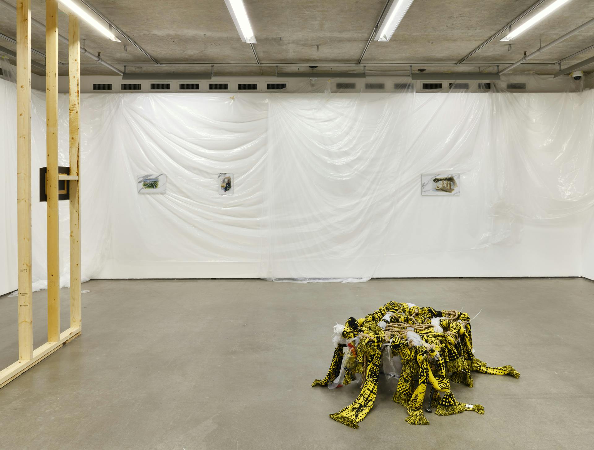 Three small paintings hang on a gallery wall draped with plastic sheetings. To the left is an unfinished wooden wall structure. A sculpture made of yellow textiles, plastic and rope sits on the floor. 