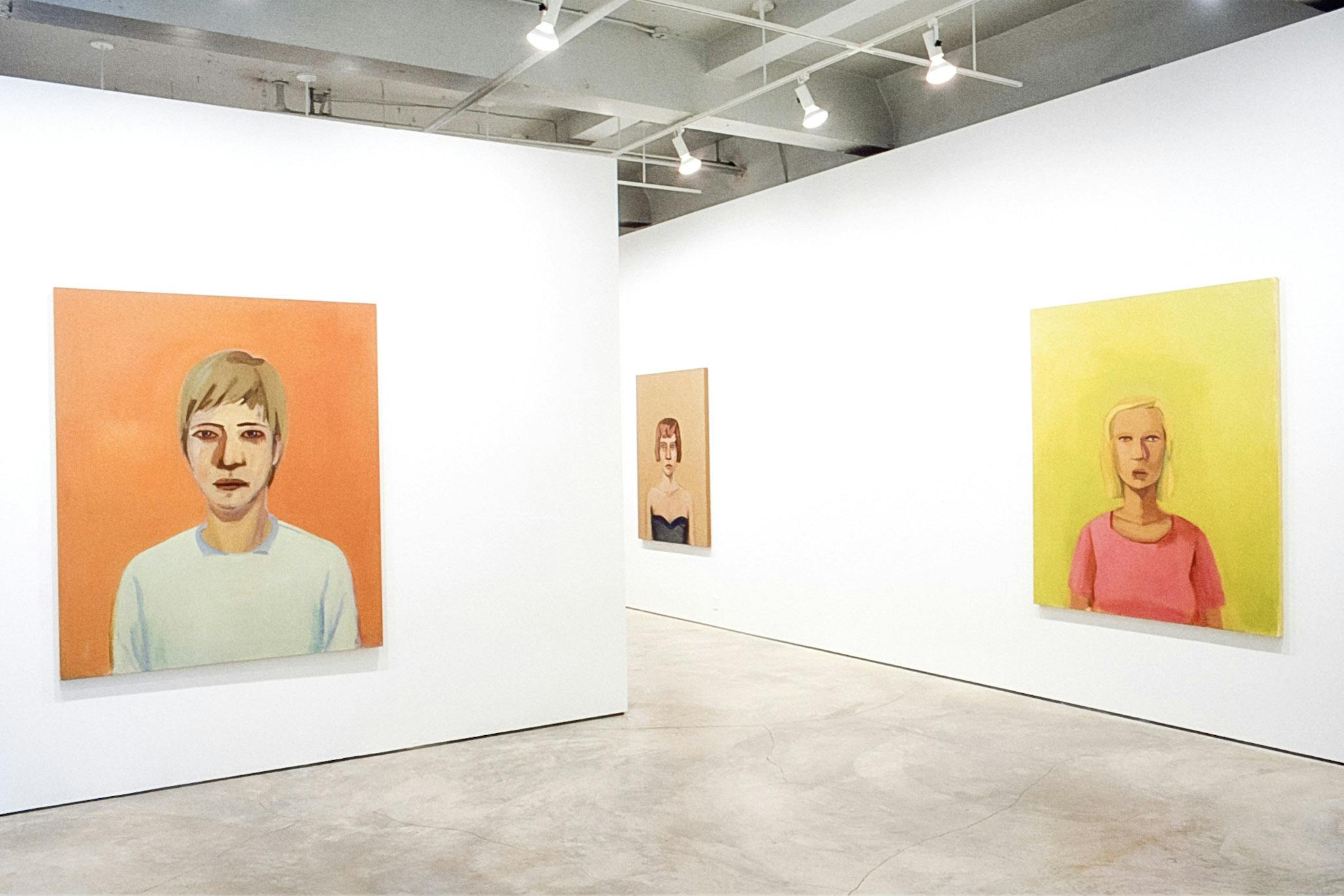 Three portrait paintings are installed on the gallery walls. The one in the front is a short-haired person standing in an orange background, wearing a white shirt and looking straight at the viewer. 