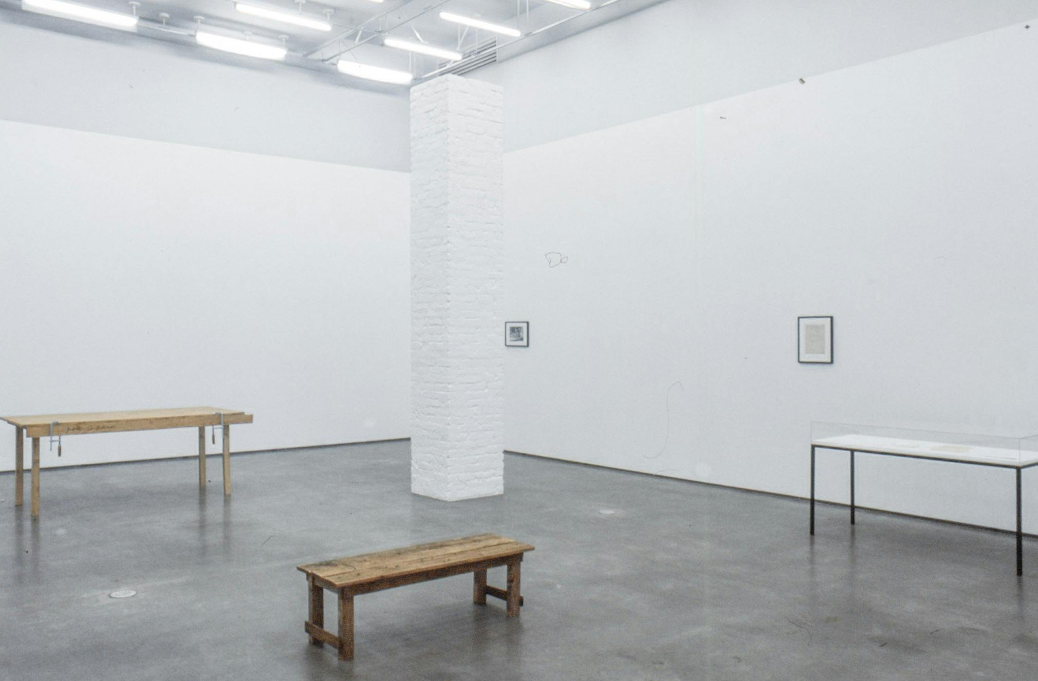 An installation image of Kirsten Pieroth’s art exhibition. At least 7 meters tall white square pillar stands in a gallery. The pillar does not reach the gallery ceiling. Several text-based and photography works are mounted on walls. 