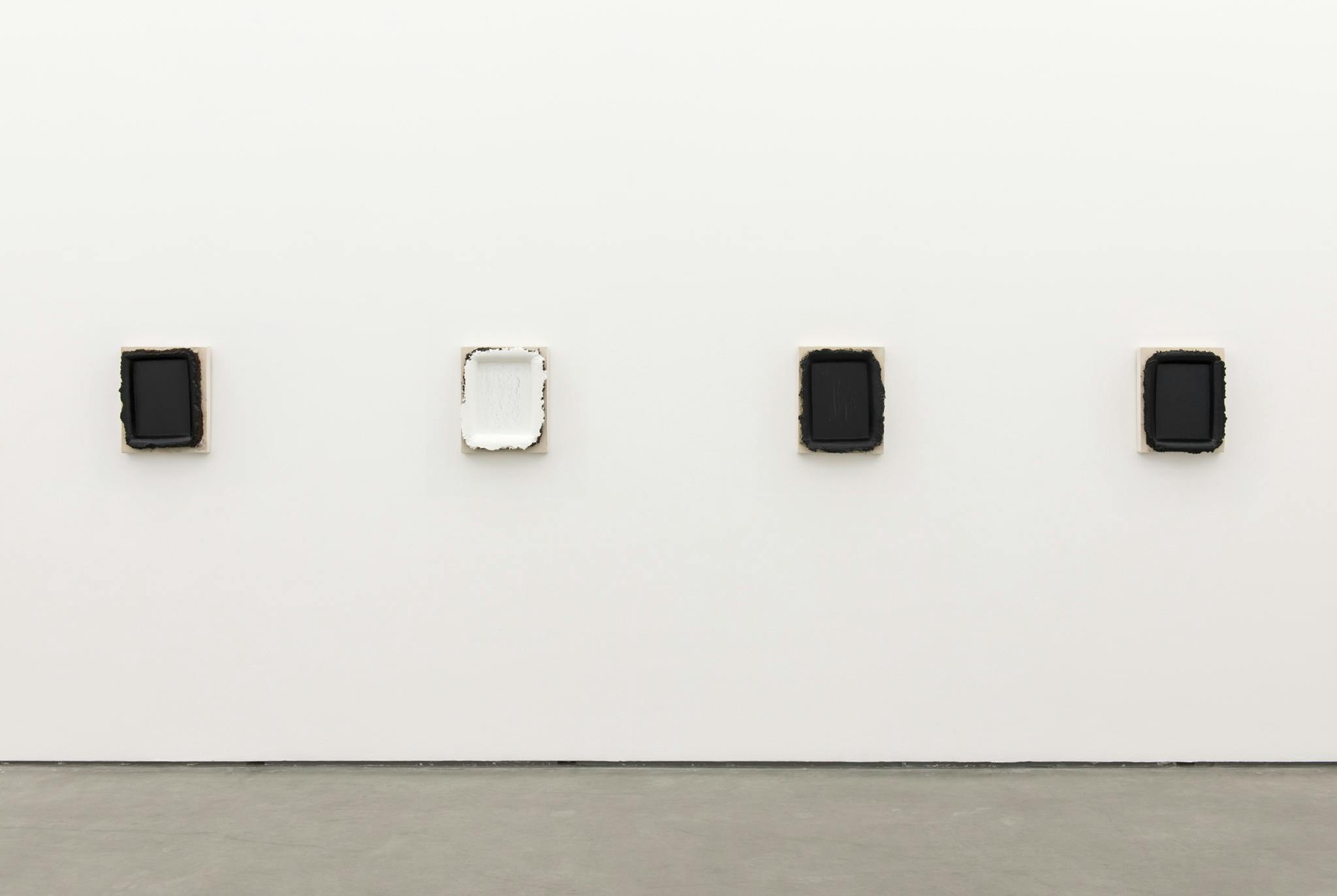 Four paintings hang on the wall of a gallery space. Three of them are black and one of them is white. The minimalist, rectangular paintings take on a sculptural form with thick layers of oil paint. 