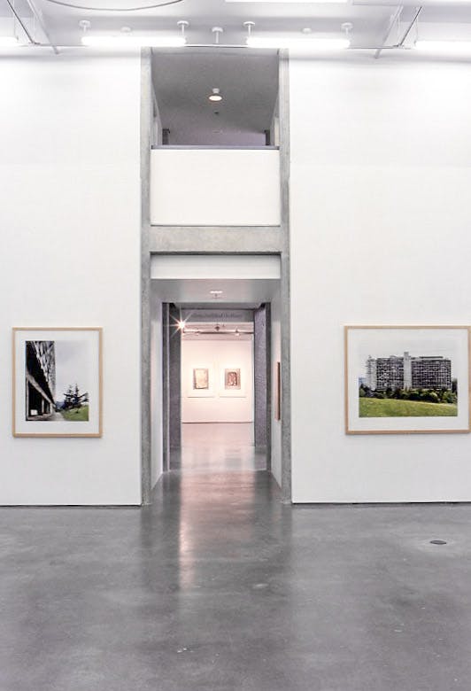 An entryway into a gallery space with concrete floors. On either side of the entry, there is a photo in a wooden frame. The photos show 2 different large buildings with large green fields beside them. 