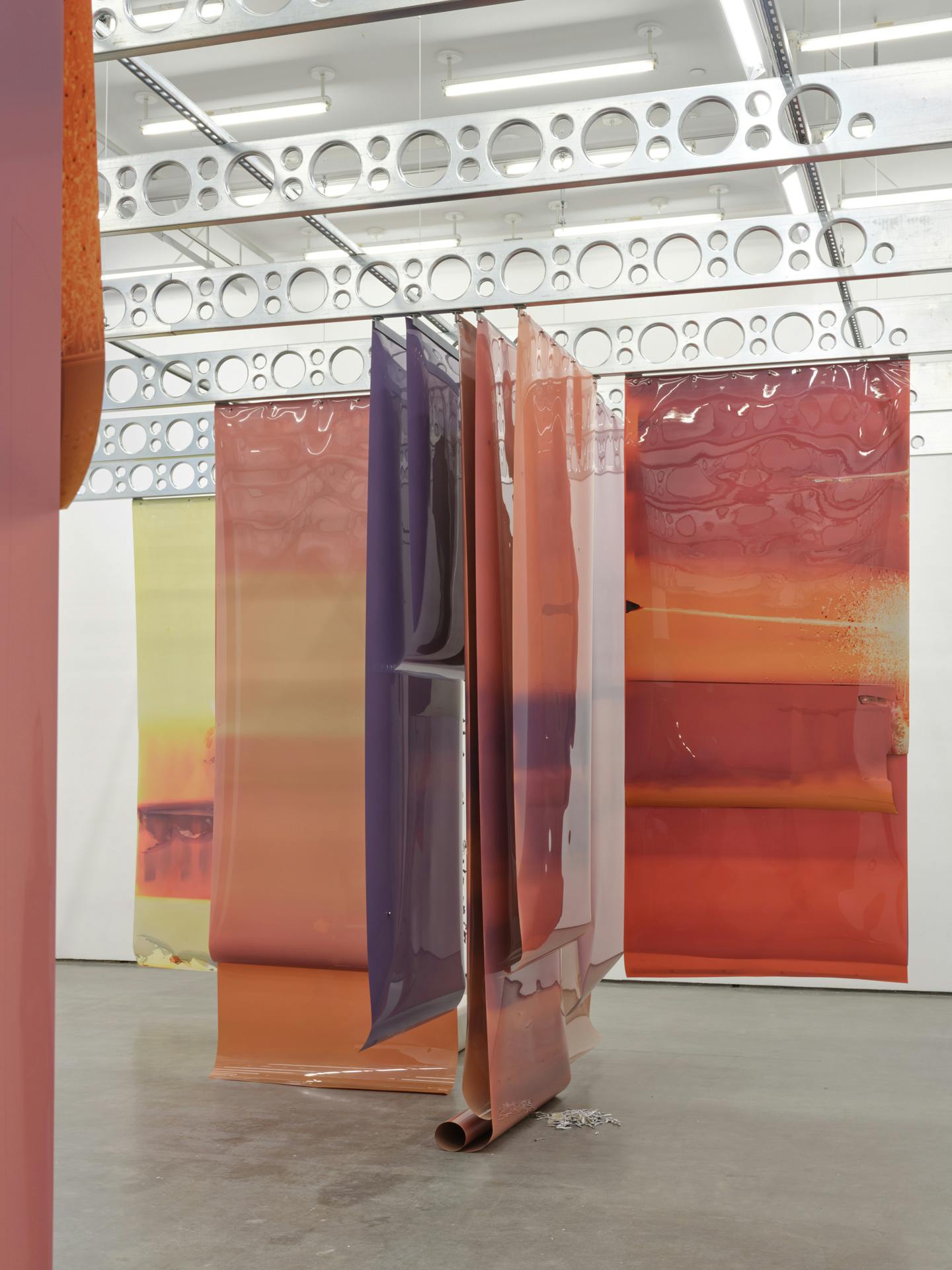 Long earth tone sheets of film of various colours are suspended from industrial steel ceiling beams. The edge of one sheet is visible along the side of the image.