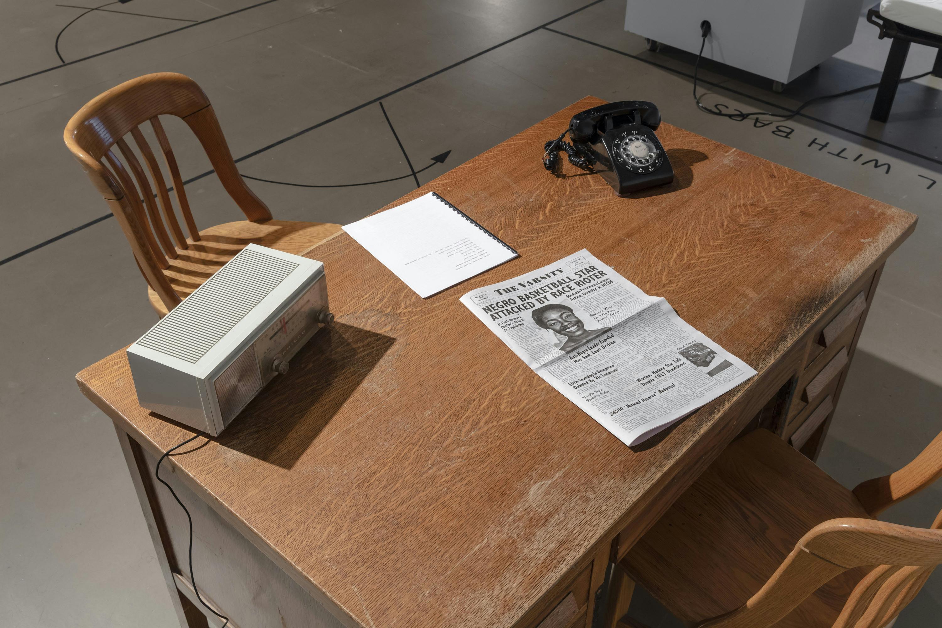 A detail of installation work by Deanna Bowen. On a table with two chairs, a rotary phone, a radio, newspaper, and a note are placed. 