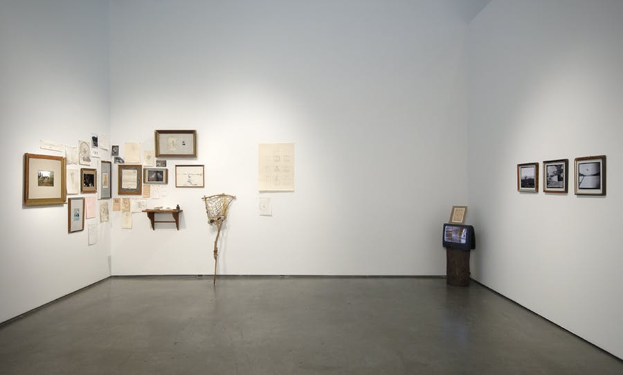 On the left corner wall, a salon style assortment of framed images, text, notes and objects hang on the wall of a gallery. On the right side, three images hang on the wall and a tv sits in the corner.