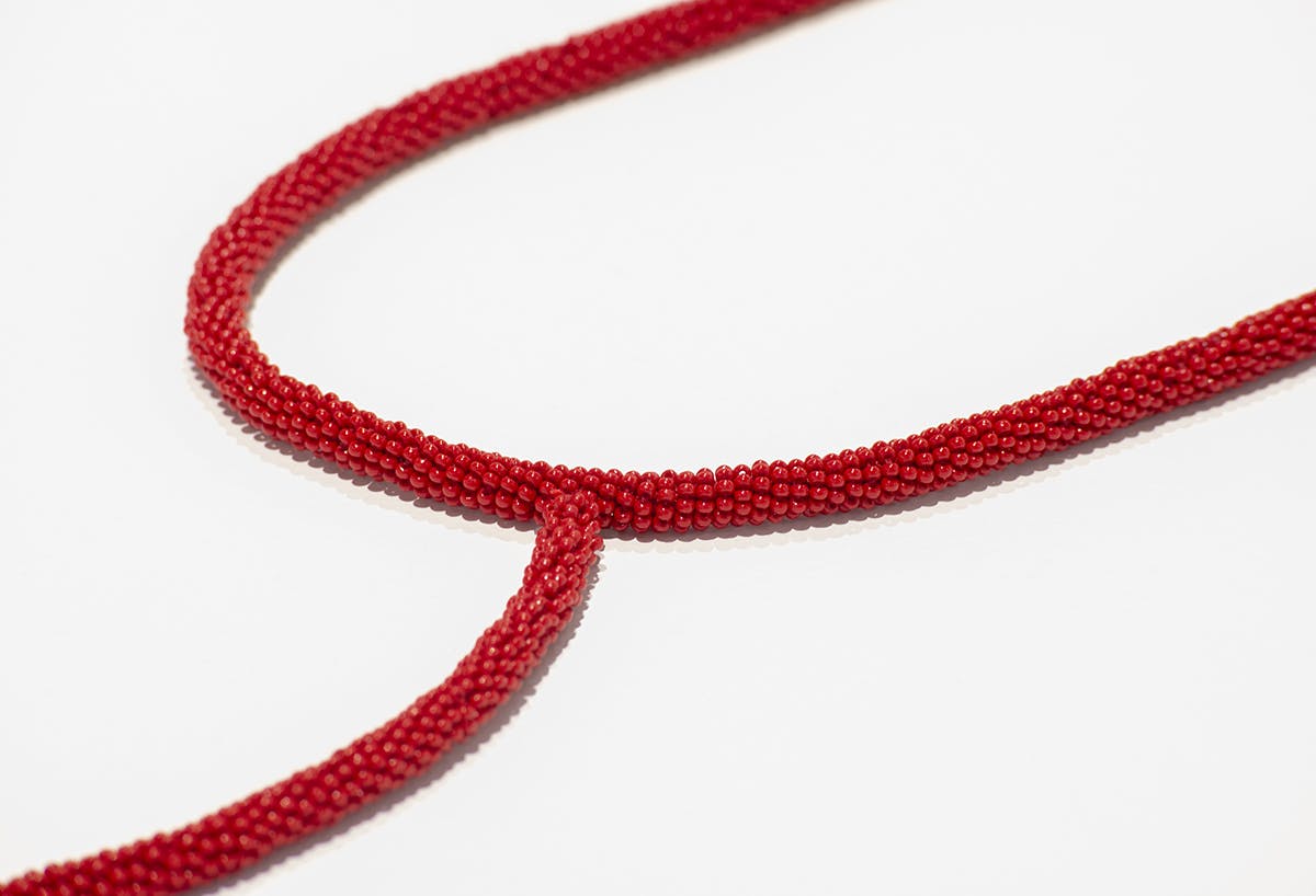 This is a close-up view of Faye HeavyShield’s beaded sculpture. It is a long unbound cord, which has been covered entirely in red beads. The surface of each bead reflects the gallery light.