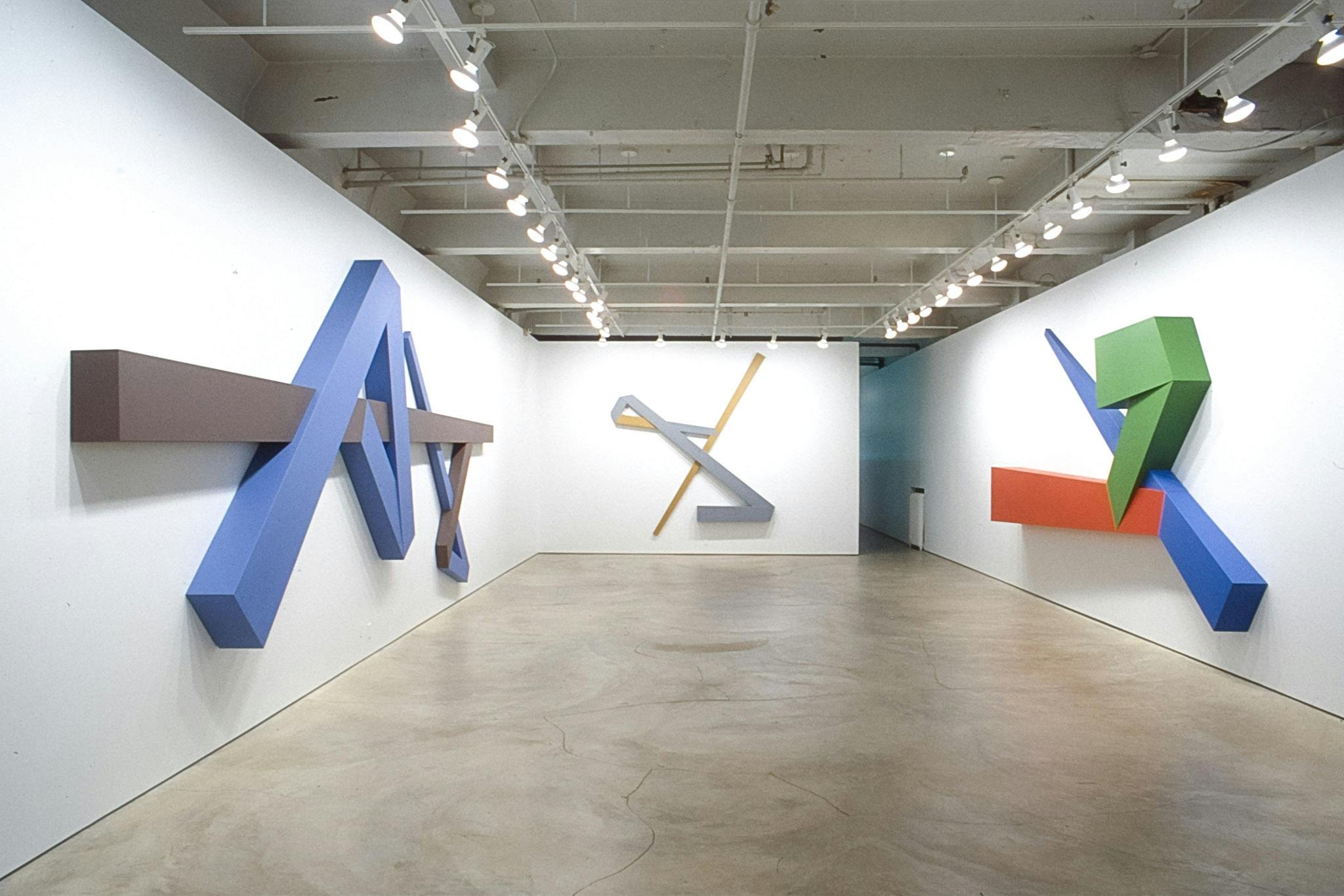 Three large-sized geometric sculptures are mounted on the gallery walls. The one on the left resembles a capital letter A in blue, and the middle one resembles a flipped over character Z in grey. 
