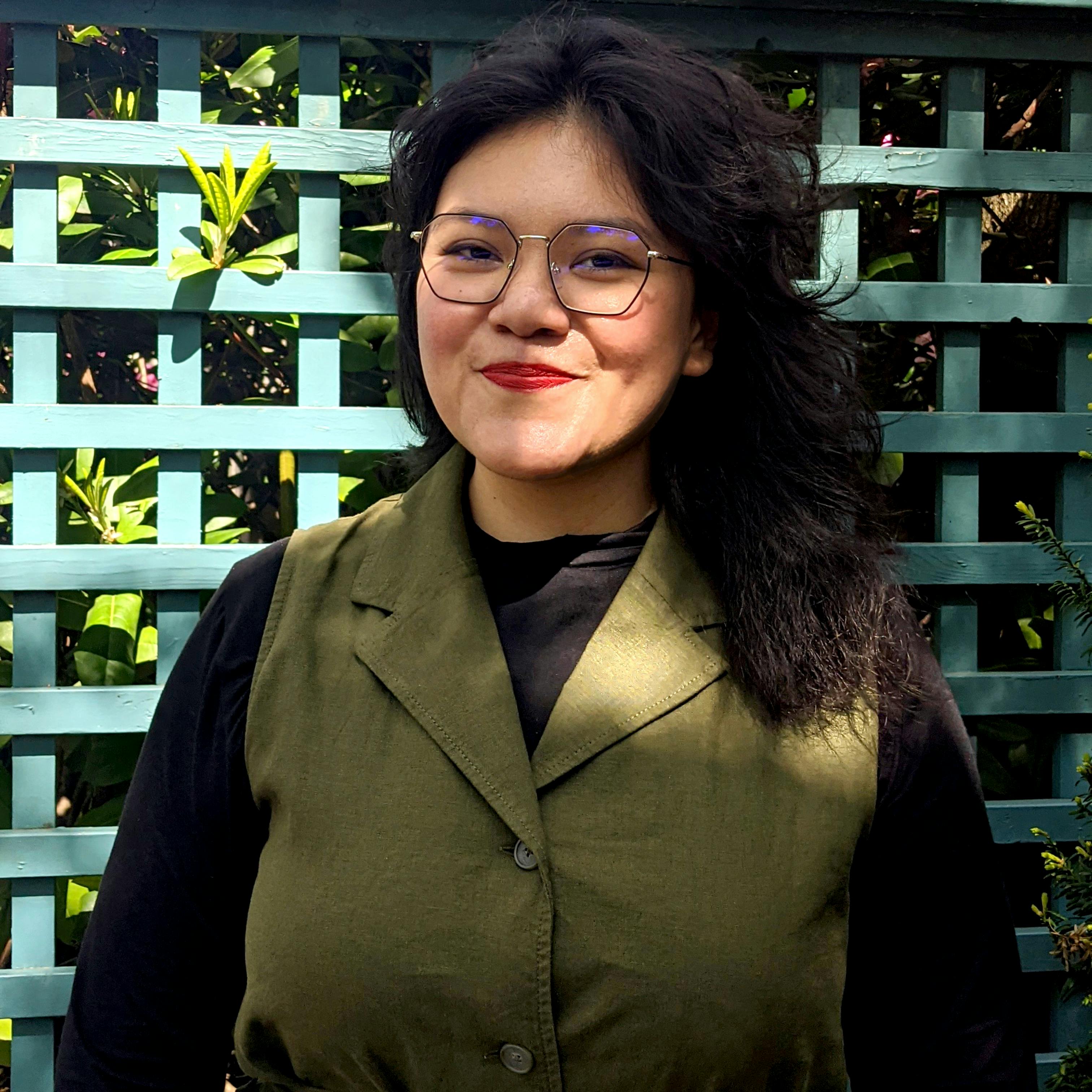 A portrait of Faune Ybarra. Faune is smiling, wearing a green vest and glasses. Behind her is a blue lattice with green plants peeking through.