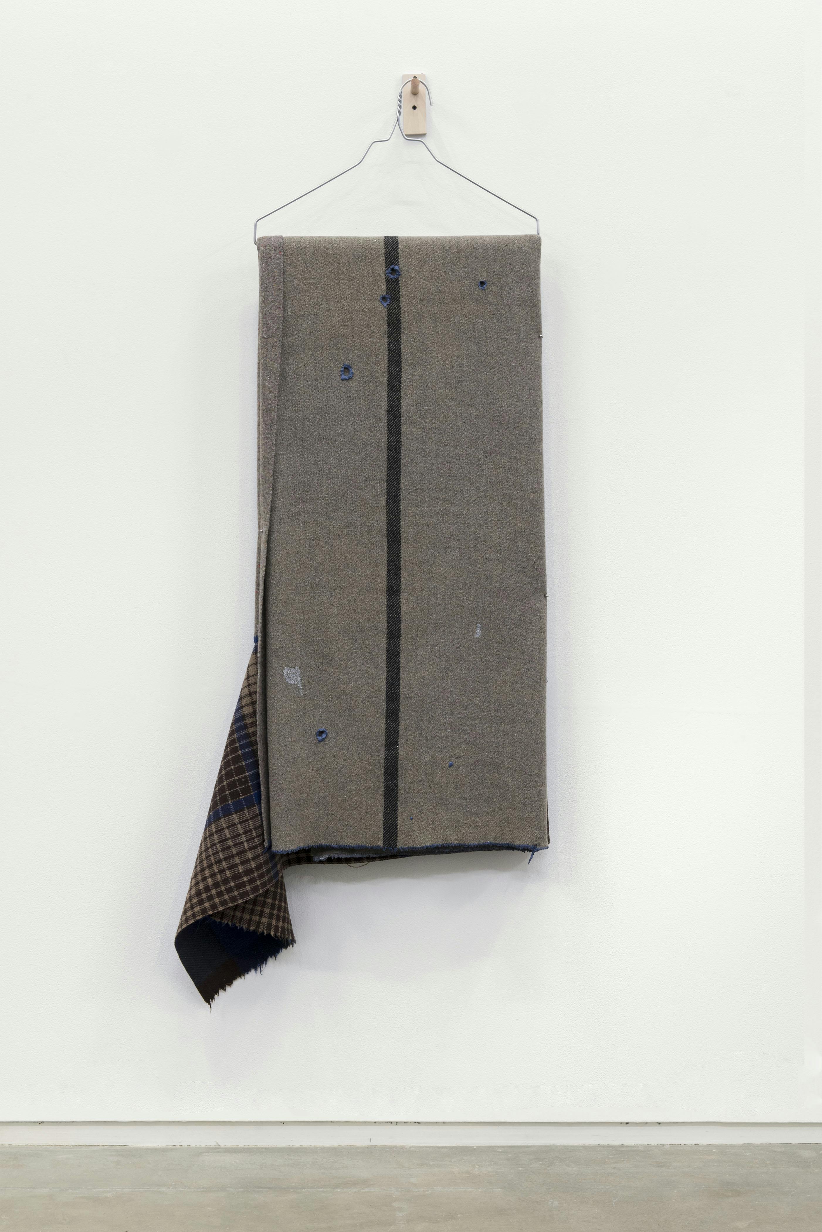 A grey textile with a vertical stripe down the middle is draped on a wire-hanger which hangs on a gallery wall.