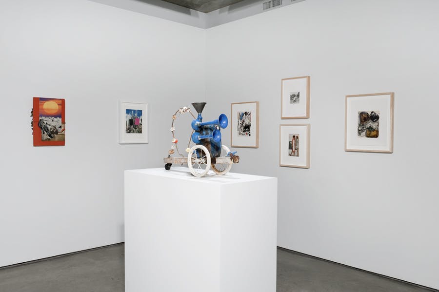 Various artworks are installed in the corner of a gallery. Framed drawings and photo-collage pieces are mounted on the walls behind a small-scale multimedia sculpture on a pedestal.