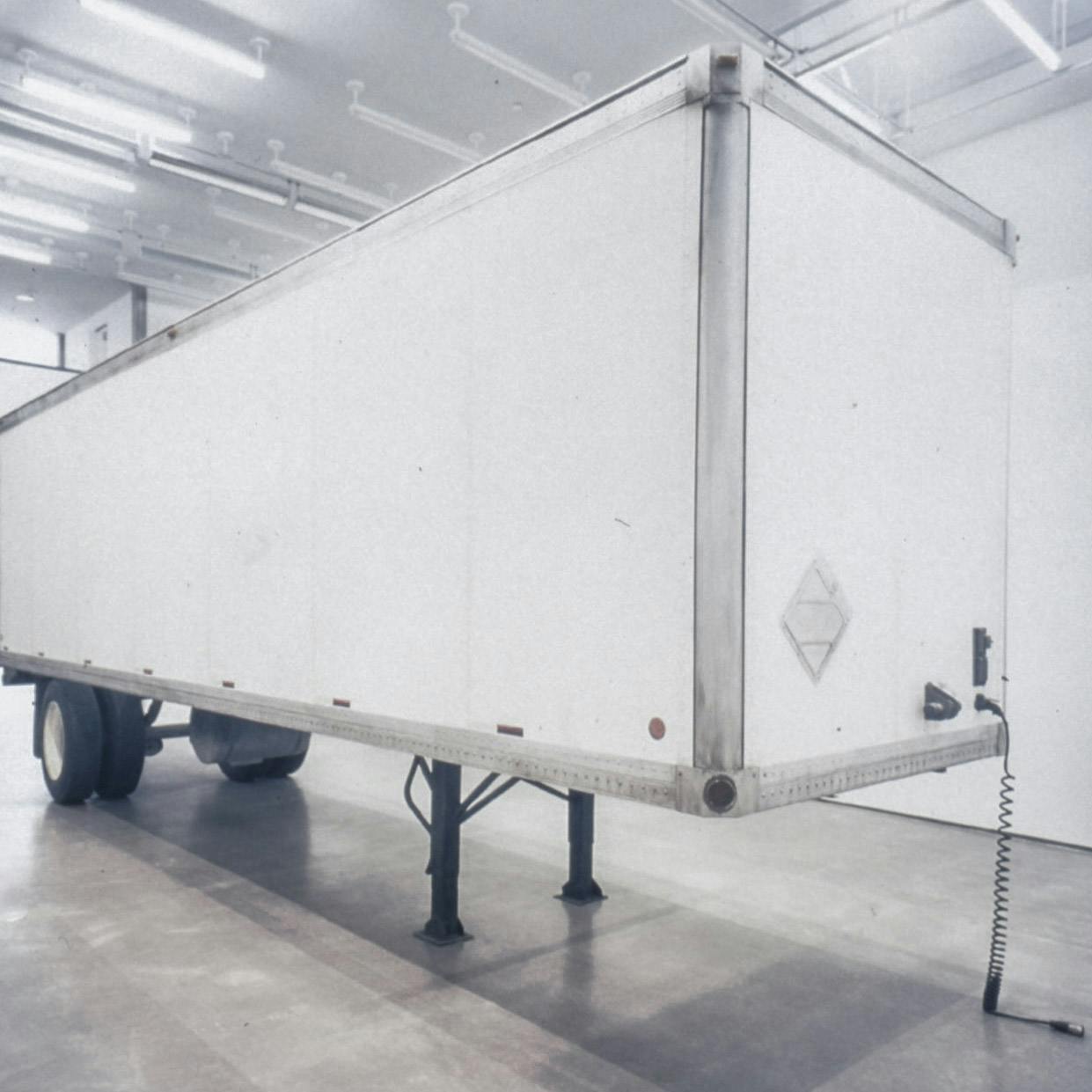 This is a side view of a white semi truck trailer installed in a gallery space. Two beams support the structure where the trailer attaches to a truck.