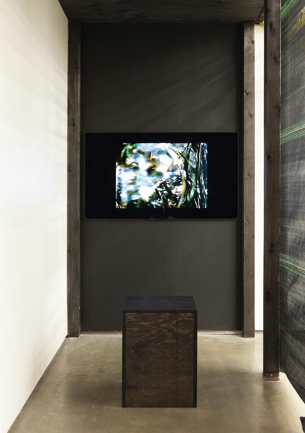 A small narrow space with a monitor, mounted on a black wall. On the screen there is an abstract blue and white image. In front there is a box seat made out of dark brown stained wood. 