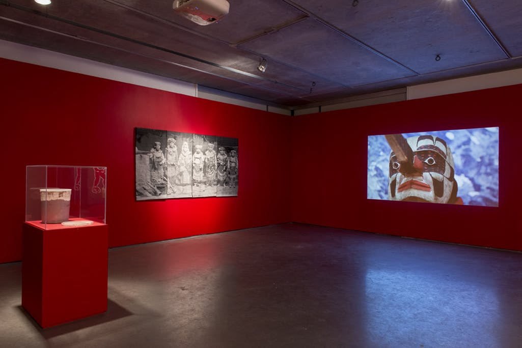 A video work by Krista Belle Stewart projected on a red gallery wall. The video shows a person wearing a carved wooden mask. A black and white photograph of children is installed on the other wall. 