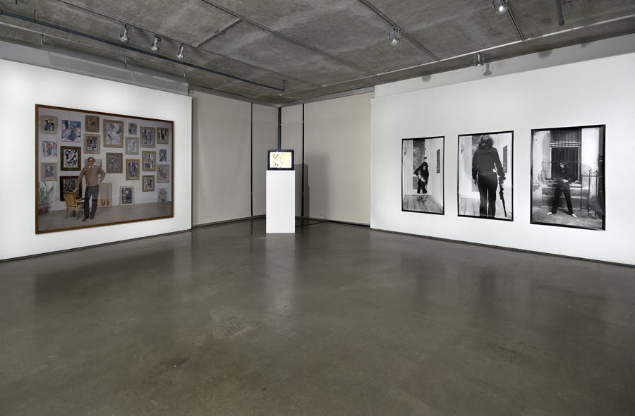 A tv on a plinth in the corner of a gallery. Large scale photographs hang on either side. One photo shows a person standing in front of a wall of paintings, the other photos are black and white.