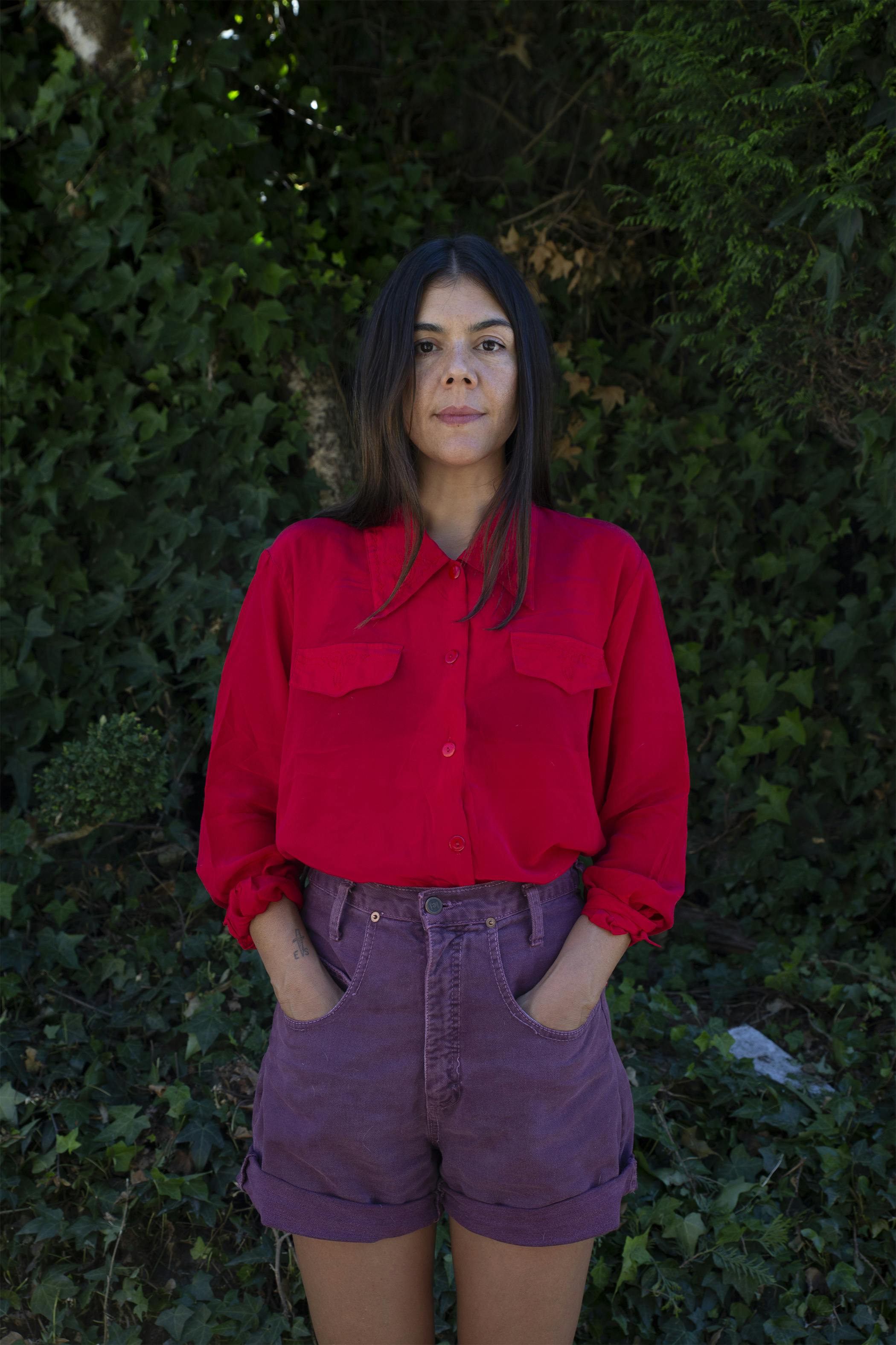 A portrait photograph of Gabrielle L’Hirondelle Hill standing in front of a bush. Hill is wearing a red shirt and purple bottoms. 