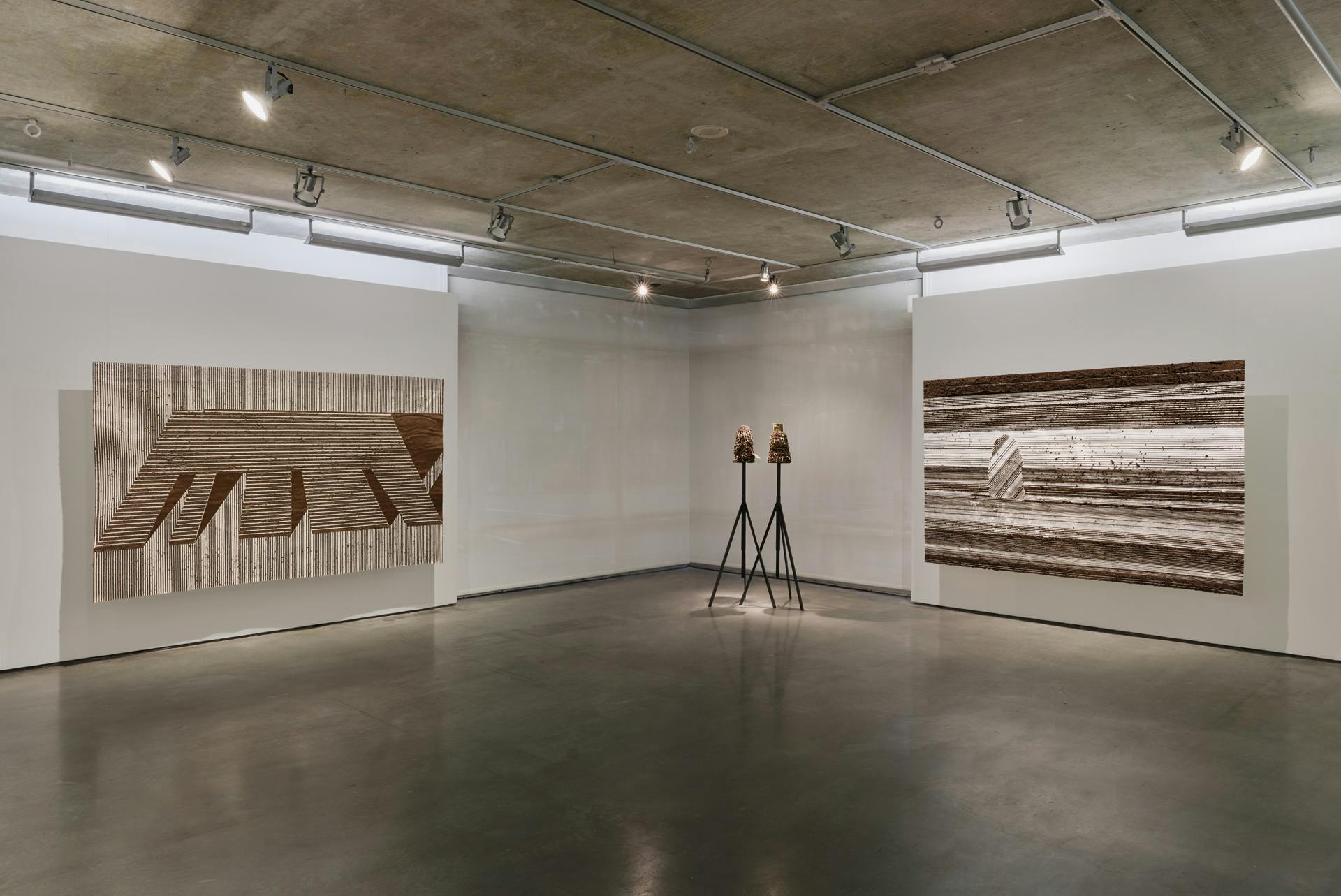 Three large scale drawings made of adobe mud and paper are suspended against the corner walls of a gallery space. In the middle sits a sculpture consisting of two mound-like forms on tripod stands.