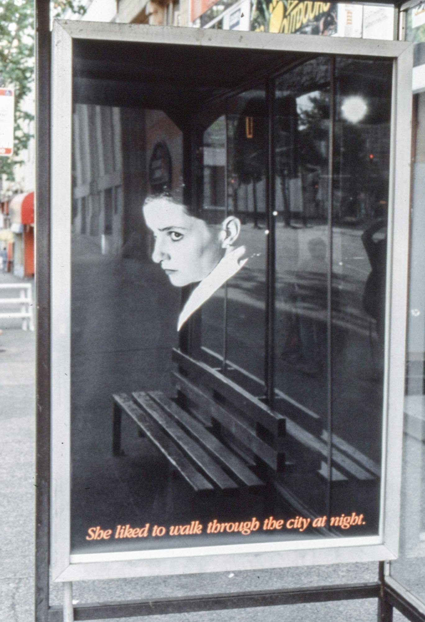 A dark photo in a bus shelter shows a person with makeup in a suit looking to the side dramatically. Orange text at the bottom of the image reads "She liked to walk through the city at night."