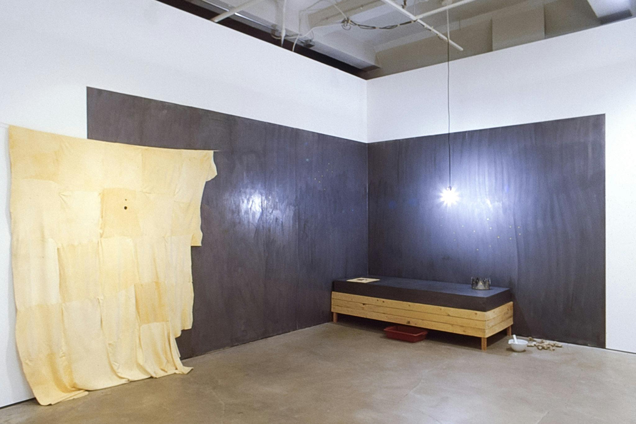 A corner of the gallery is painted in black. An old yellow blanket is hung at the end of the black-painted area. A low wooden bed frame with a black matless is placed in front of those black walls. 