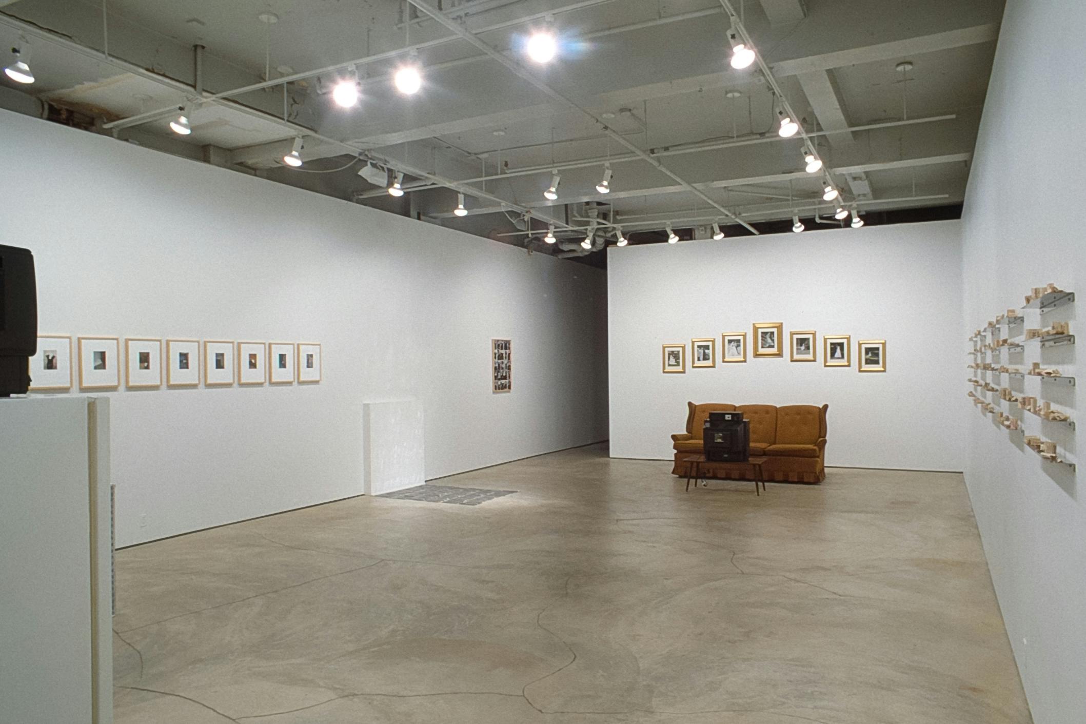 Multiple artworks are installed in the gallery. A mustard-coloured couch is placed with a CRT TV at the end of the gallery. Small-sized sculptures and pictures are mounted on the white walls.
