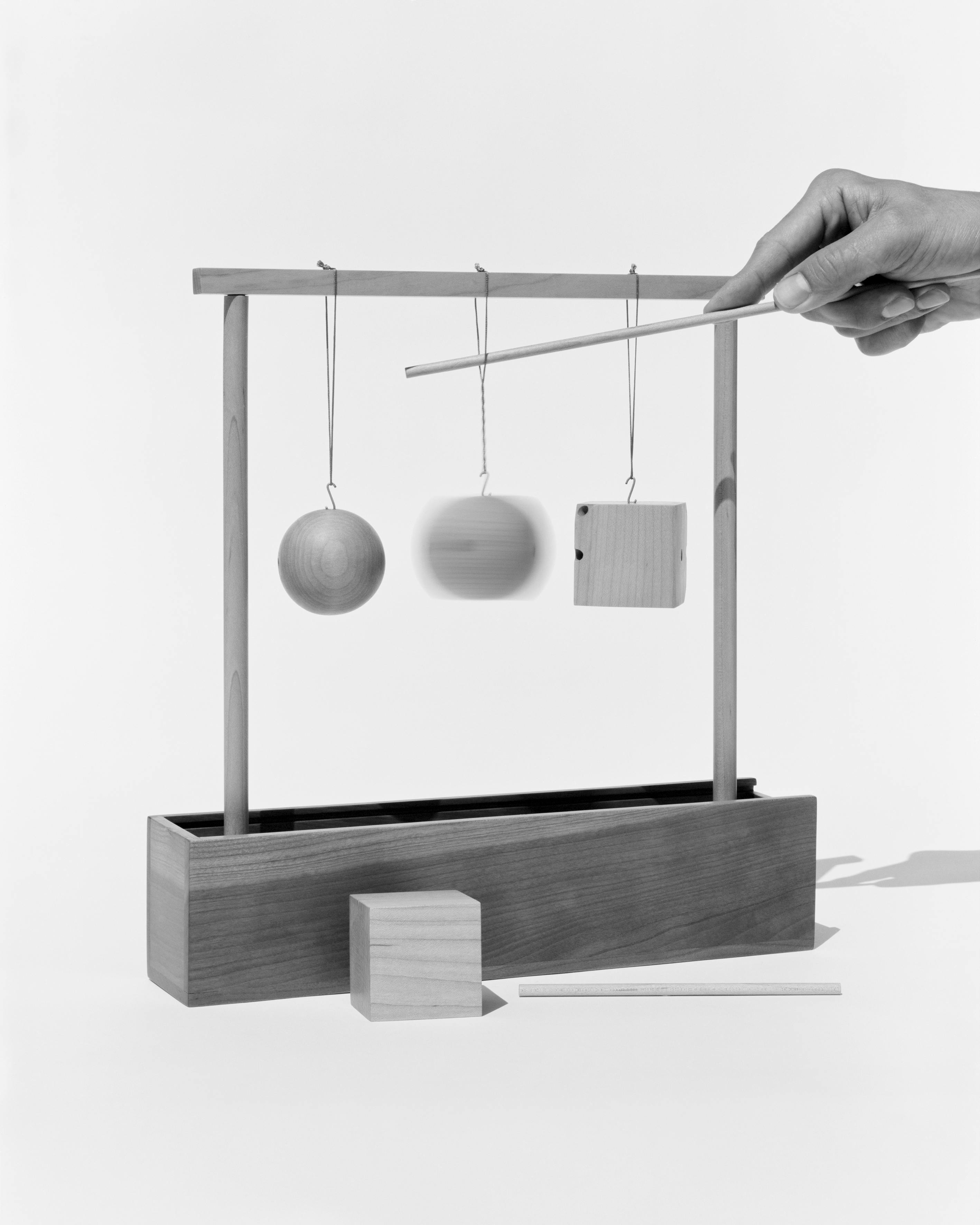 A wood object with differently-shaped blocks hanging from its simple square frame. A wood cube and dowel lay in front. A hand holding a thin wooden down reaches towards the object. 