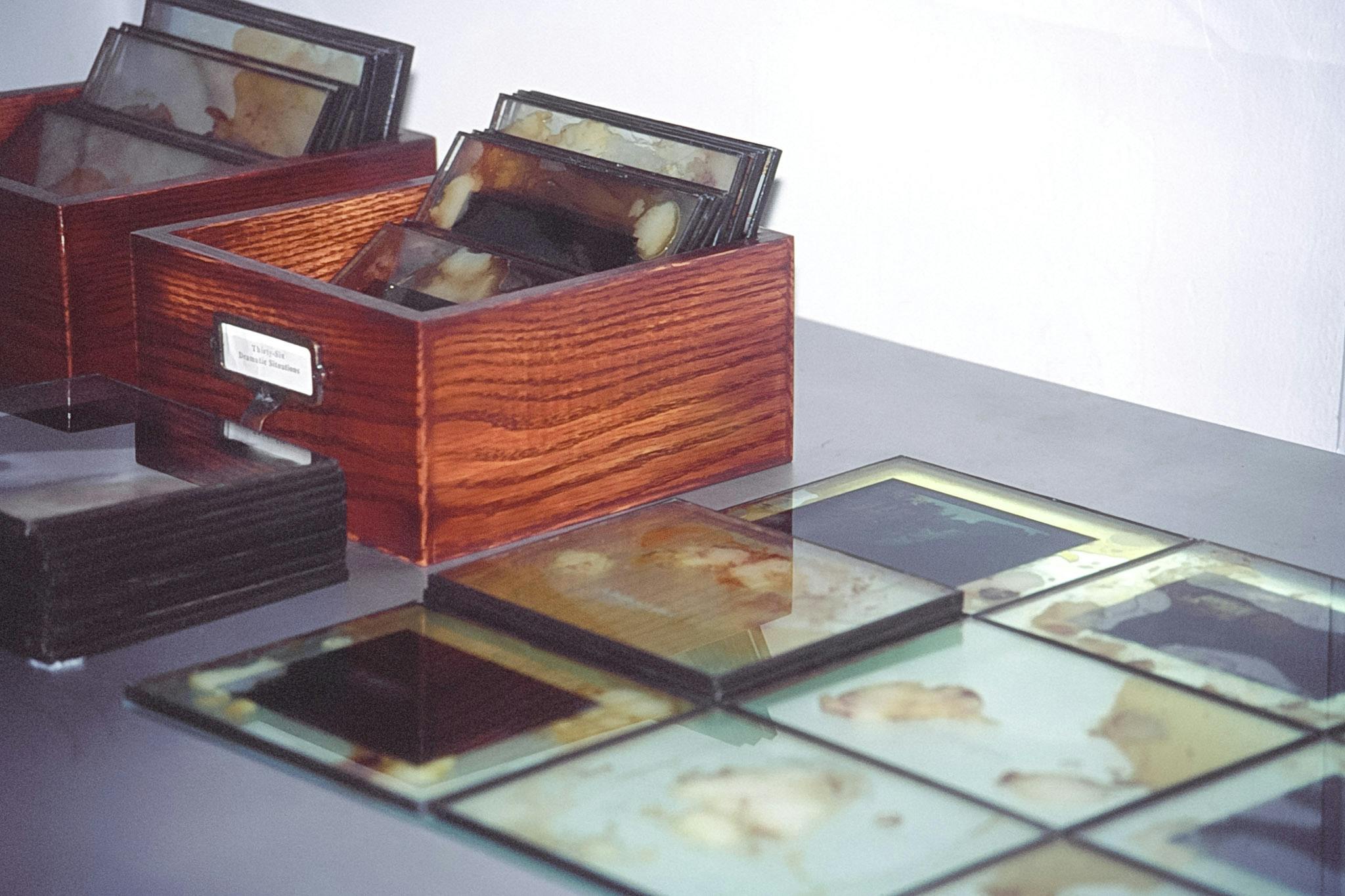 A closeup of a grey surface in a gallery. On it, two wooden boxes with several photo-transparencies in metal frames with yellow stains lay. More of these lay in front of, and beside, the boxes.