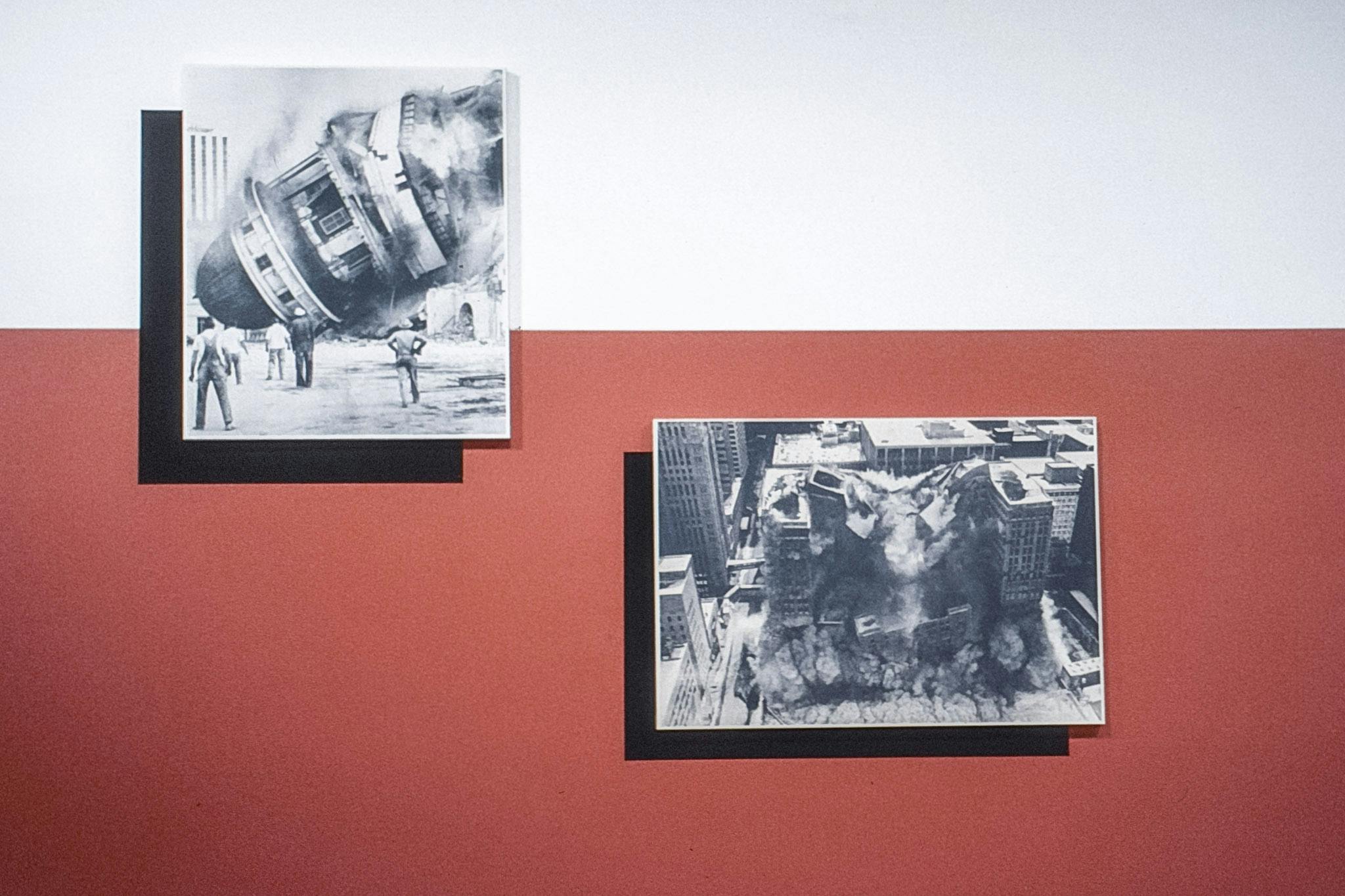 Two art works on a red and white wall. The works are black and white photos of buildings collapsing. In one, people stand, witnessing the disaster, and the other photo is from a bird's-eye view.