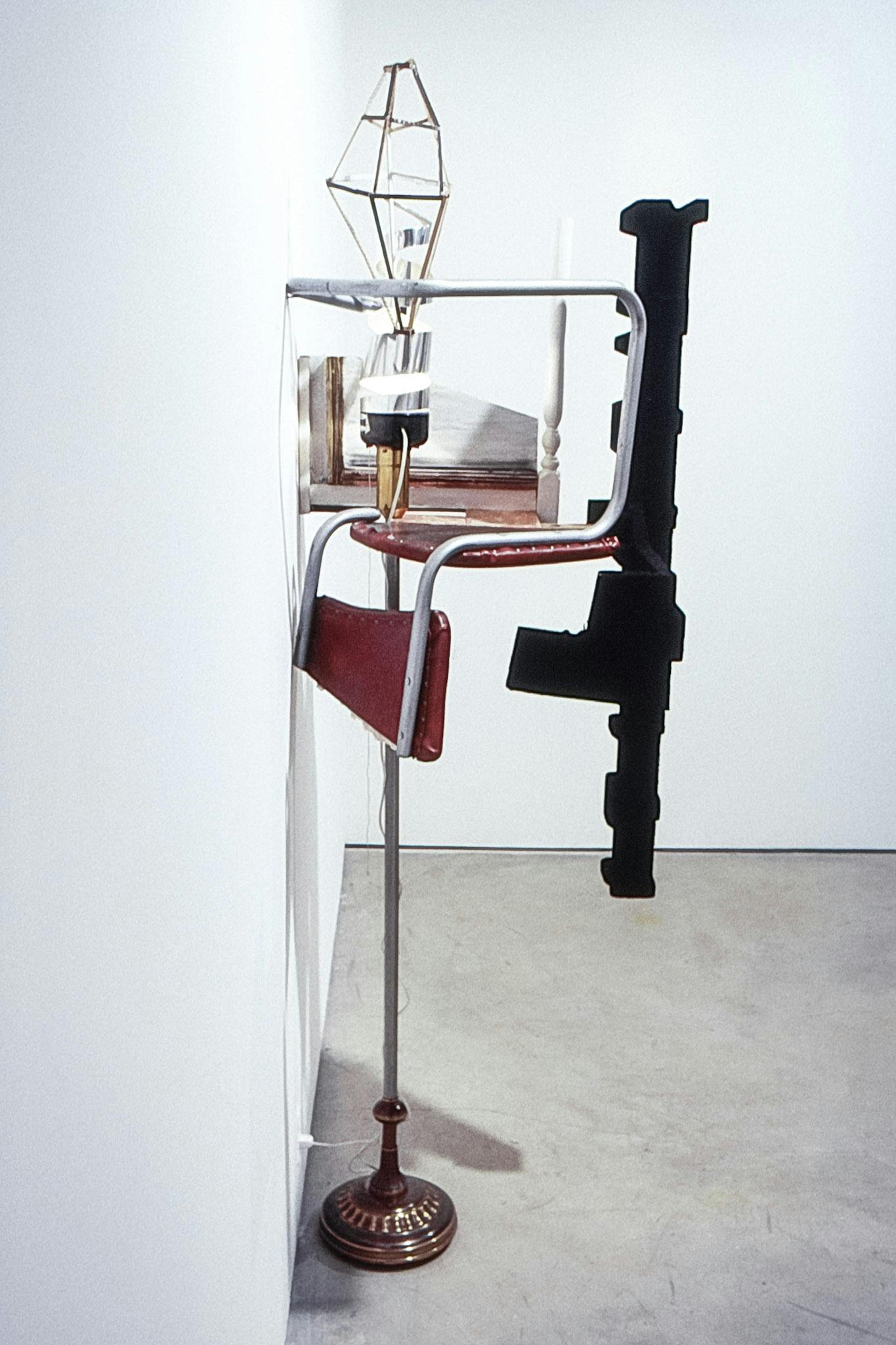 A closeup of a narrow sculpture against a wall. It is composed of different objects, including a lamp base, a bent cushioned metal chair, an upside down end table, and a long black foam form.