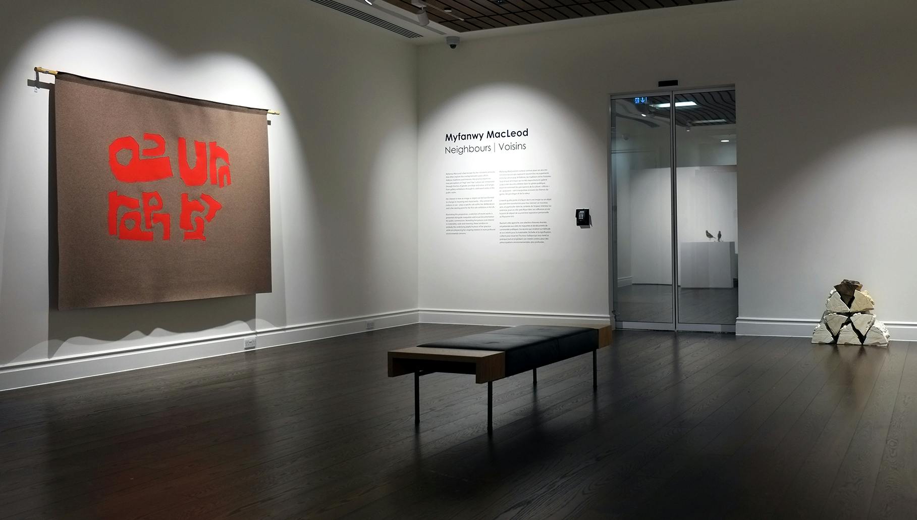 A view of Myfanwy MacLeod’s exhibition in a gallery. On the left wall, brown fabric with red shapes hangs off a curtain pole. On the right side,  a pile of plaster cast wooden logs sit on the floor.