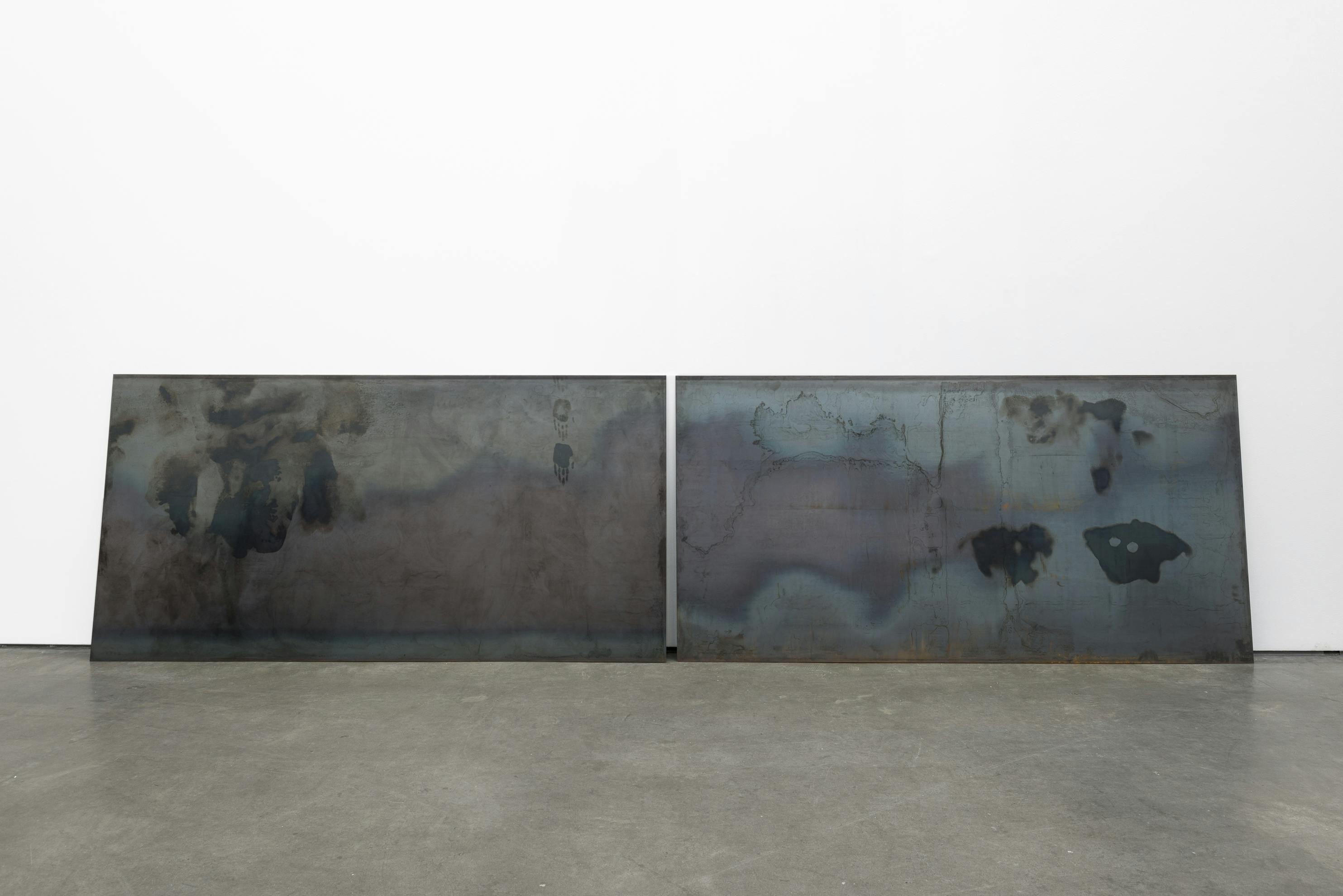 Two large rectangular steel sheets lean against a wall of a gallery space. The sheets sit horizontally on the floor and are marked with grease prints. On the left sheet two handprints are visible. 