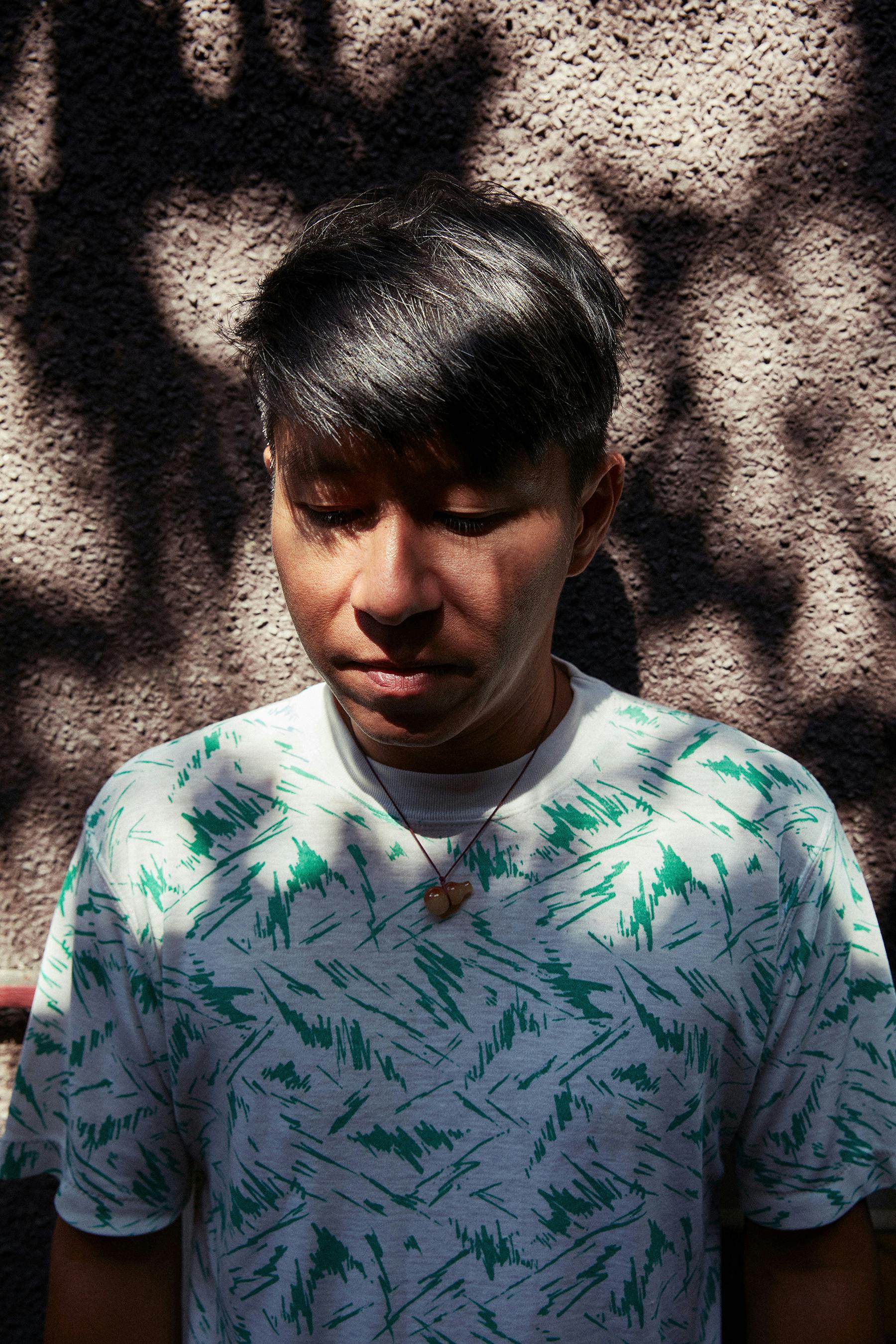 A portrait of Howie Tsui. He is standing against a wall wearing a white shirt with a scribbly green design. He is shaded by the speckled light from an unseen leafy canopy above.