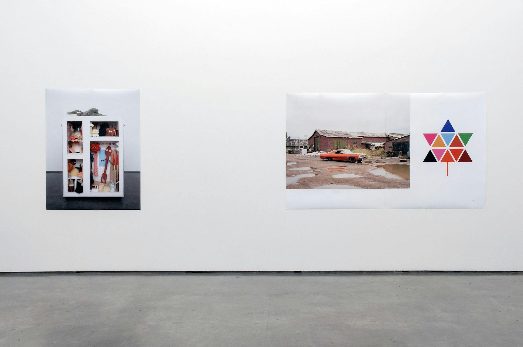 Two large-sized posters are installed on a gallery wall. The one on the left side is a photograph of a shelf filled with toys, and another poster shows countryside scenery with a car and a house. 