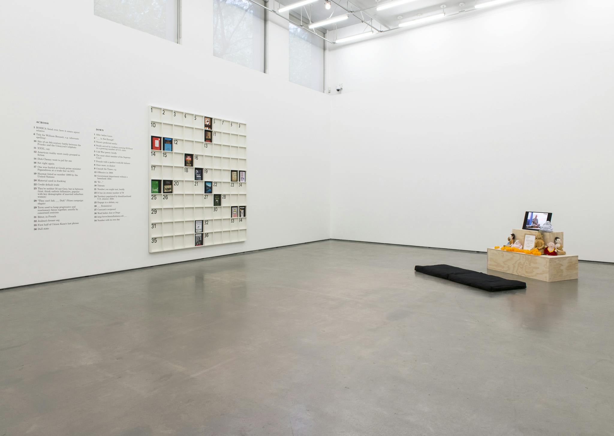A sculpture forming a large-scale crossword puzzle hangs on the wall. Across the gallery, a wooden, two-tier box displays Buddha toys and a monitor. The monitor displays video of soldiers' meditating.