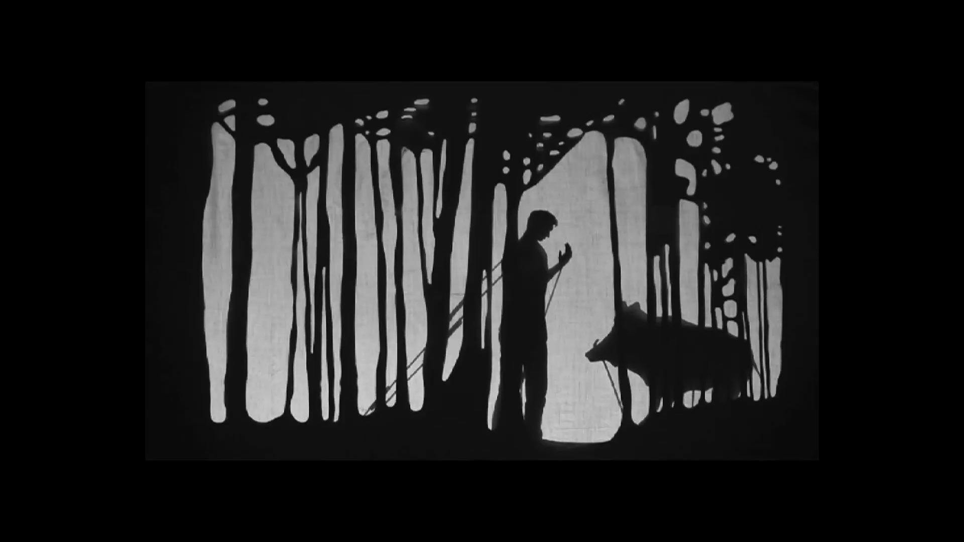 Image still from a video of shadow puppets. A figure of a man stands before a wild boar in a forest, their arm raised as if in conversation with the boar.