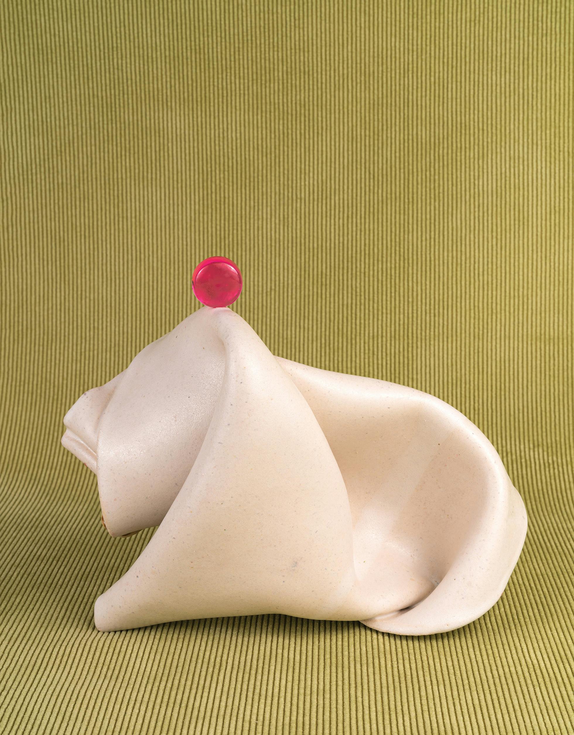A photographic work by Michelle Bui. A folded white napkin, which balances a small pink round object at its top, is placed on a green textured backdrop.