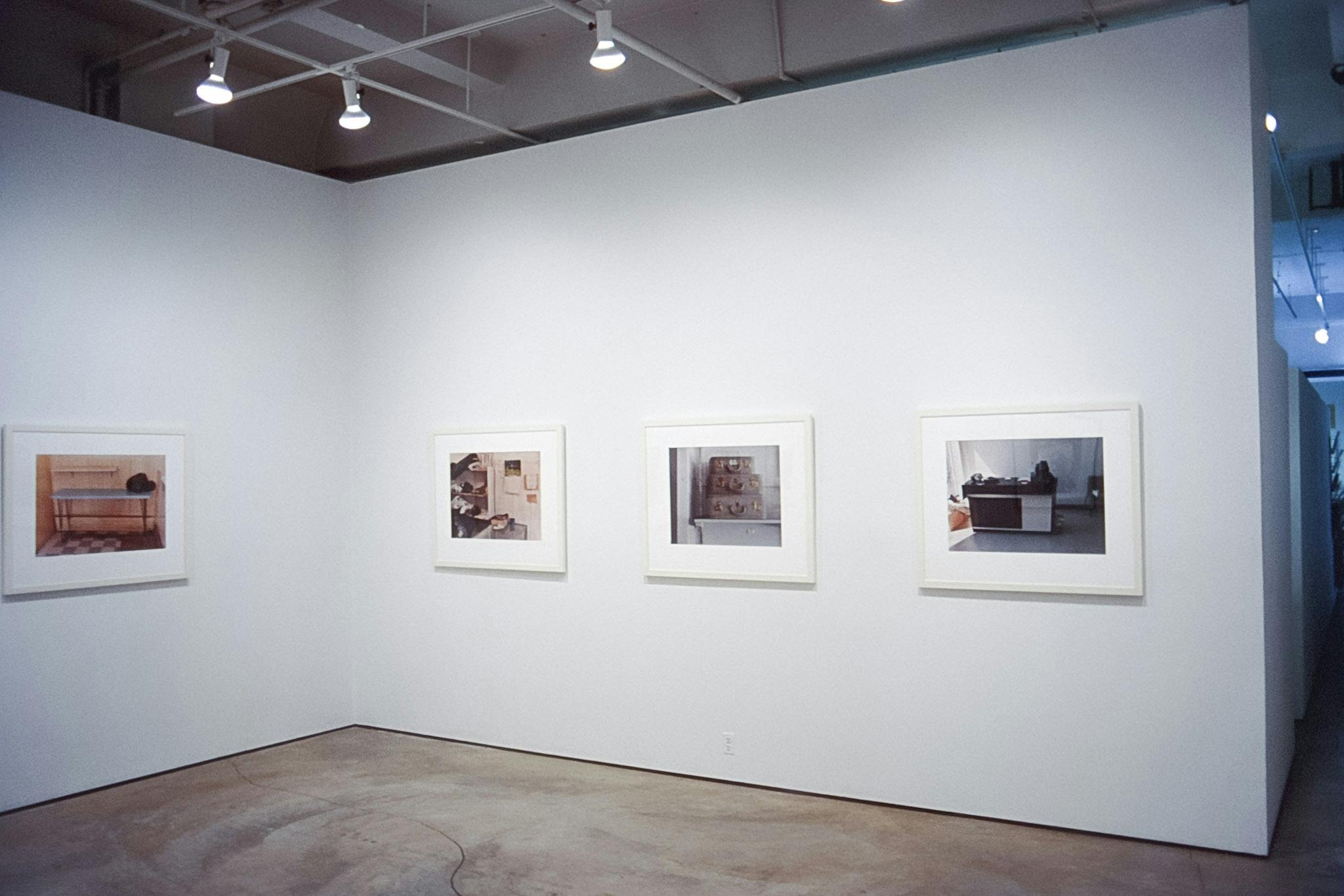 Four coloured photo pieces are mounted on the gallery walls. All of them depict items placed in white rooms. A microwave, suitcases, shelves, and benches are captured in these photographs. 