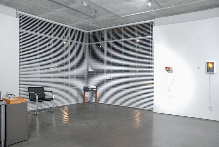 An installation image of Daniel Olson’s art exhibition at CAG. A corner of the galley is covered by glass and white window blinds. A black office chair and a projector are placed in front of these blinds.