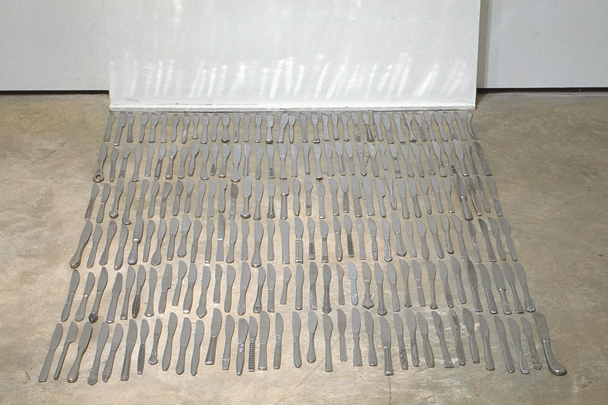 Six rows of thirty-five silver dinner knives are placed on the gallery floor. Those different sets of knives face their cutting edges to the right and point to the white wall.  