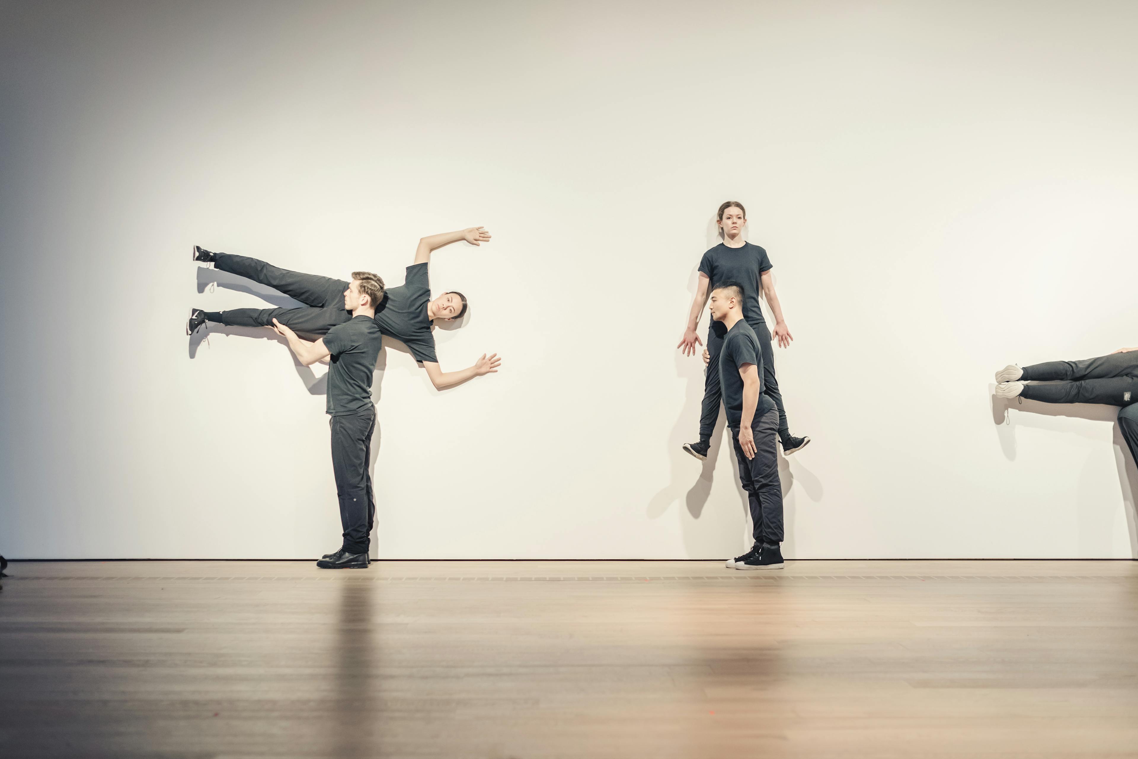 Still image of people in black t-shirts performing choreography. Three pairs of two dancers are in various poses, many leaning against the walls. 