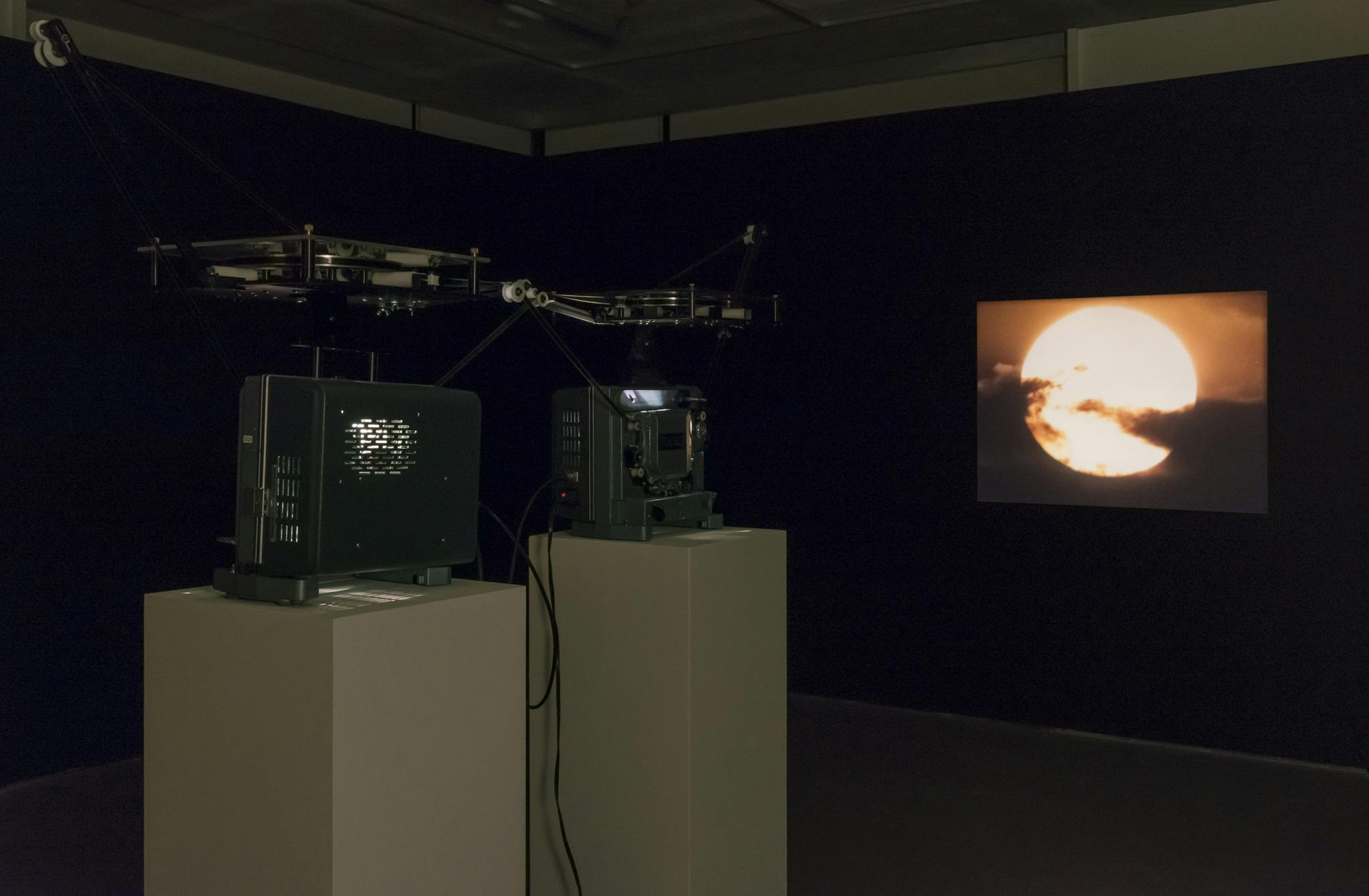 A dark gallery space with painted black walls and two projectors installed on white plinths. The projectors thread a single film between them, projecting a video of the sun. 