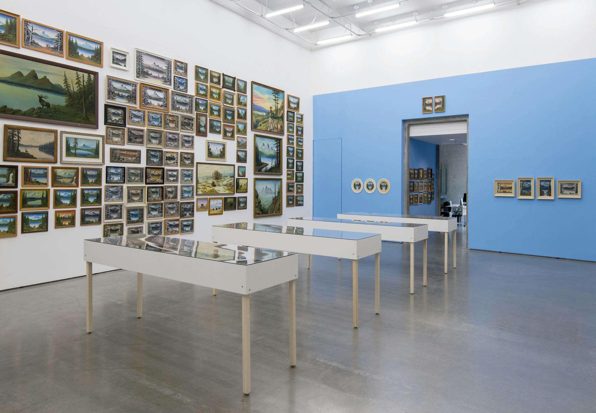 Multiple framed paintings of similar mountain landscapes cover one wall of a gallery. An adjacent wall is painted bright blue and nine more landscape paintings hang. Four vitrines sit in the center.
