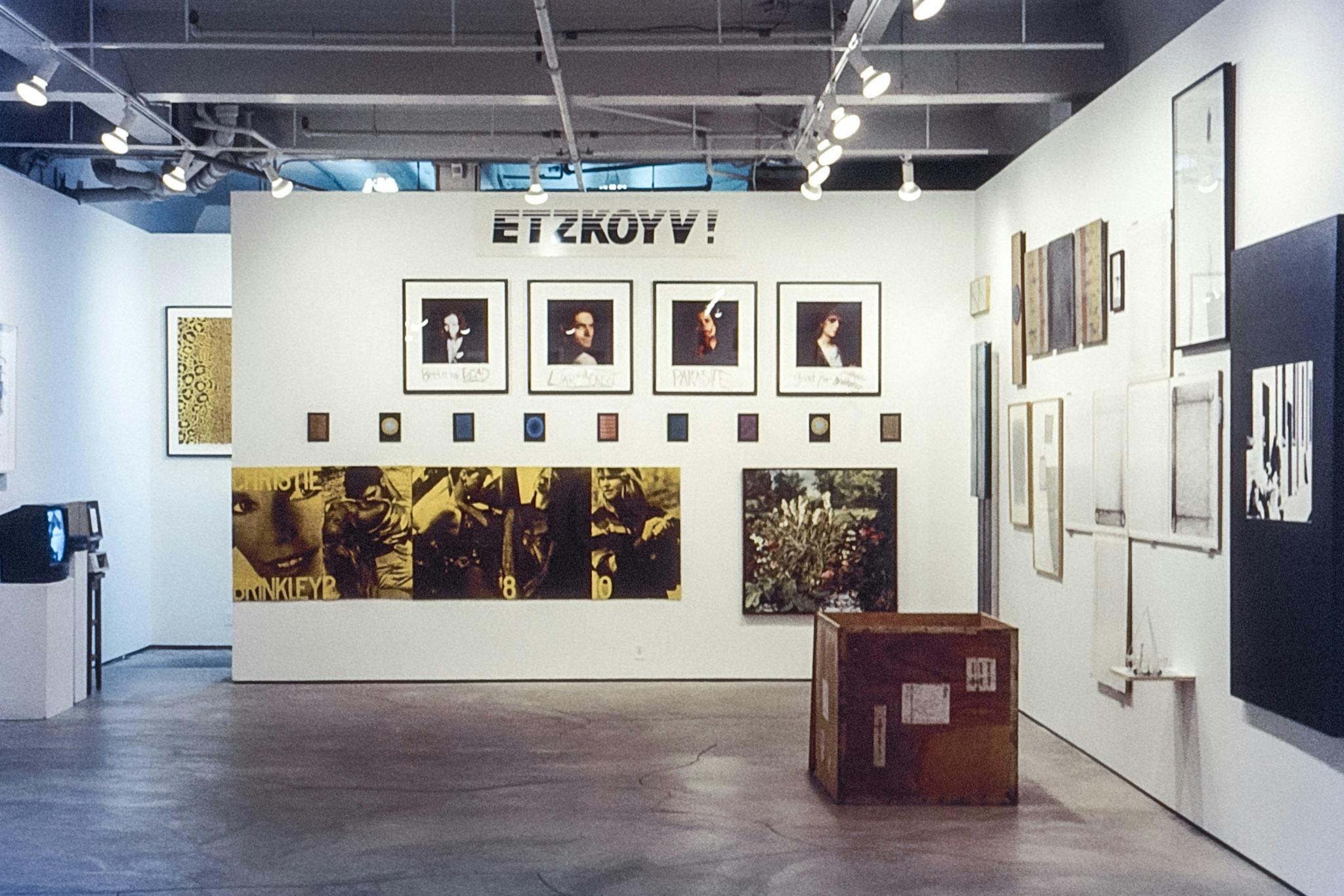 A photo of various artworks installed close to each other in the gallery. In the middle wall, the capital letter “ETZKOYV!” is placed on the top, and placed beneath are photographs of diverse people.