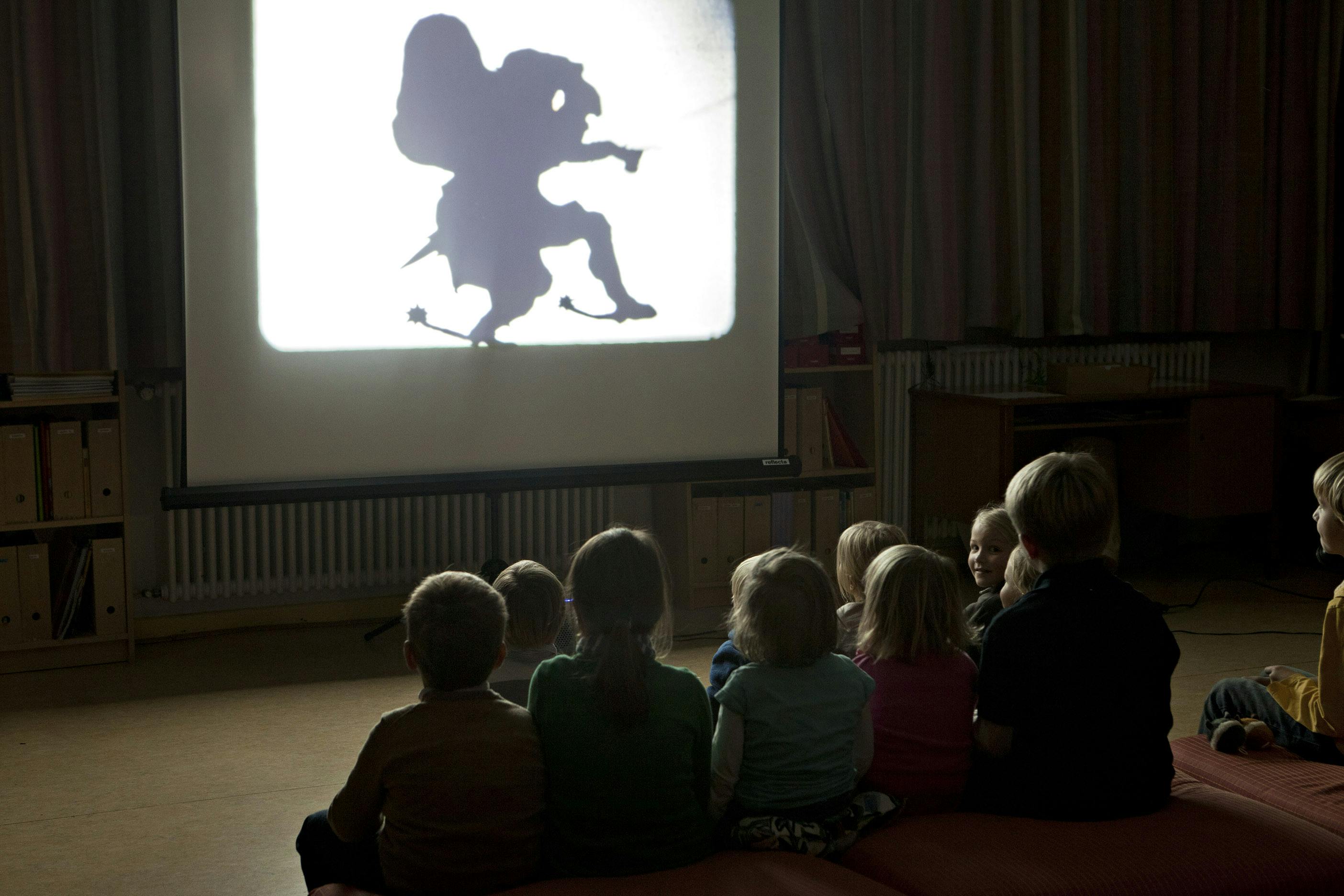 A group of young children sit in a darkened room in front of a screen watching a projected video. The video depicts a shadow puppet-like, human figure tip-toeing across the screen carrying a large sack.