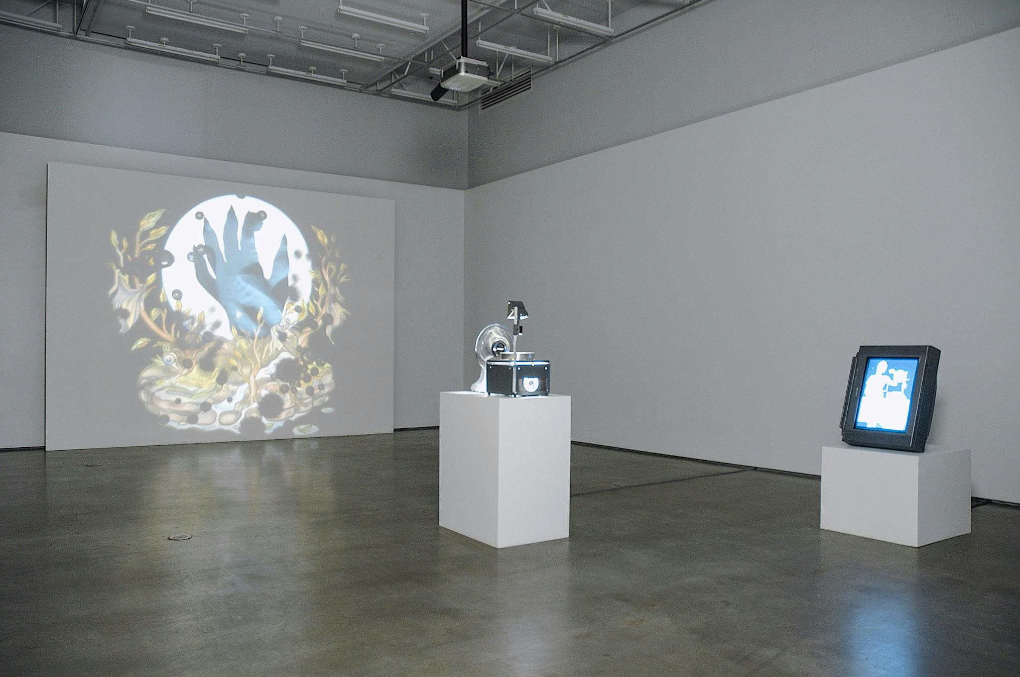 An installation image of artworks in a gallery. A projector placed in the middle of the room project an illustration of a blue hand on a far wall. On the right side of the projector, an illustration of a person is displayed on a TV screen.