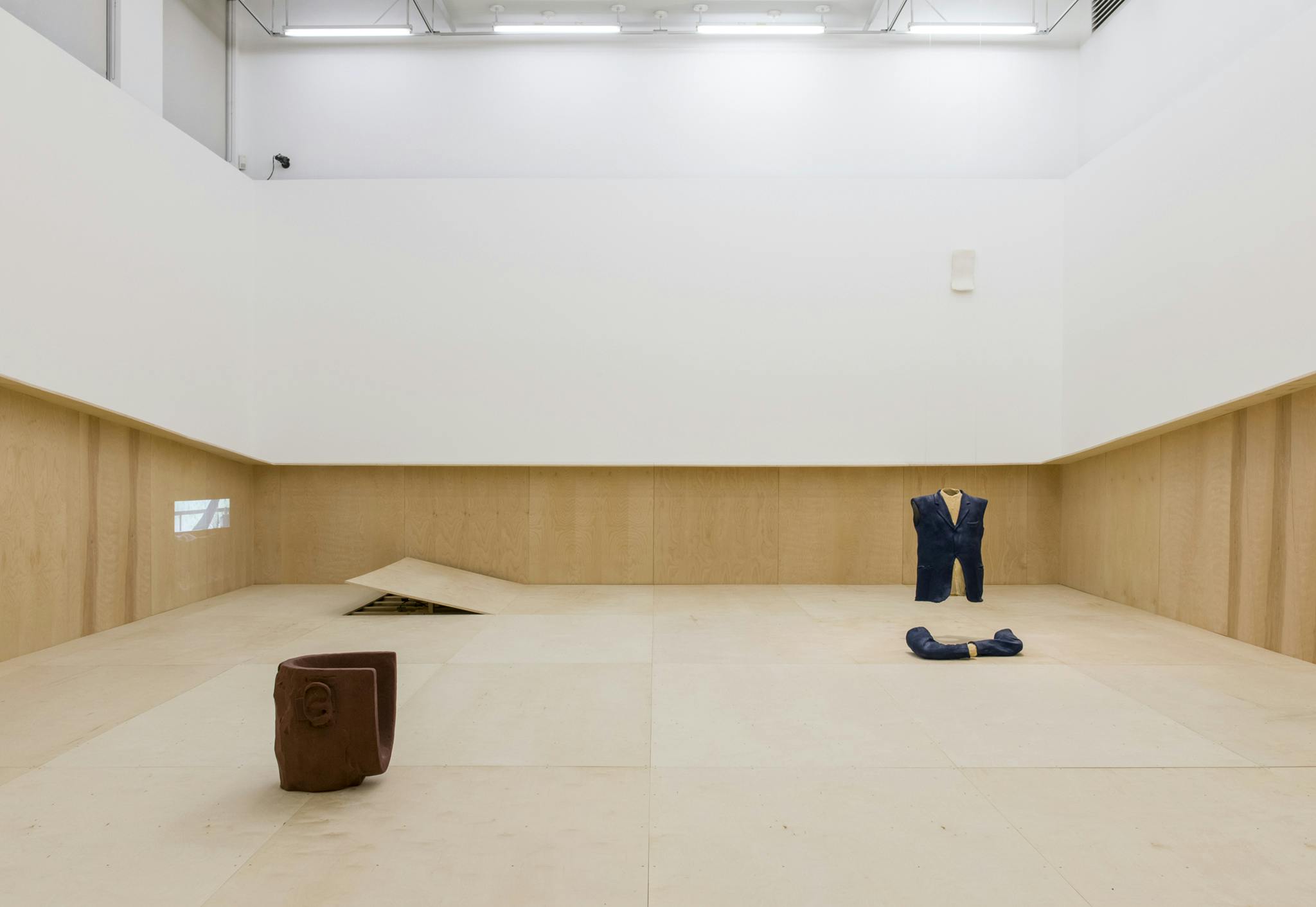 Large plywood sheets panel the floor and half the walls of a gallery. A sculpture of a torso in a blue jacket is installed on the right, a projection and a brown, chair-like sculpture on the left. 